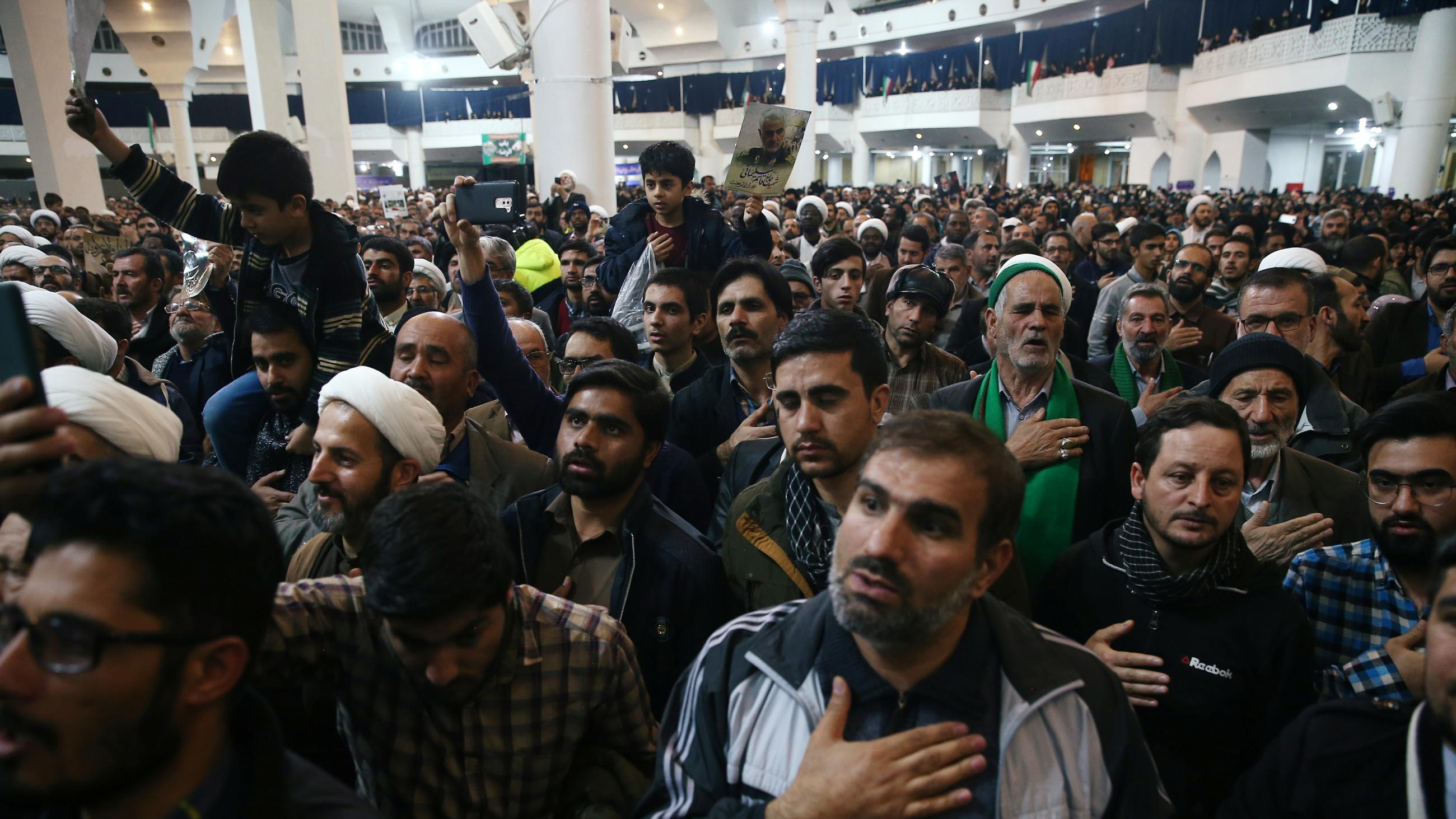 The picture shows the interior of a huge space packed with people standing. Some have their eyes open, and others have their eyes closed, presumably praying. Some hold their hand over their heart. Some hold cell phones up. A few young boys are on top of the shoulders of grown men. 