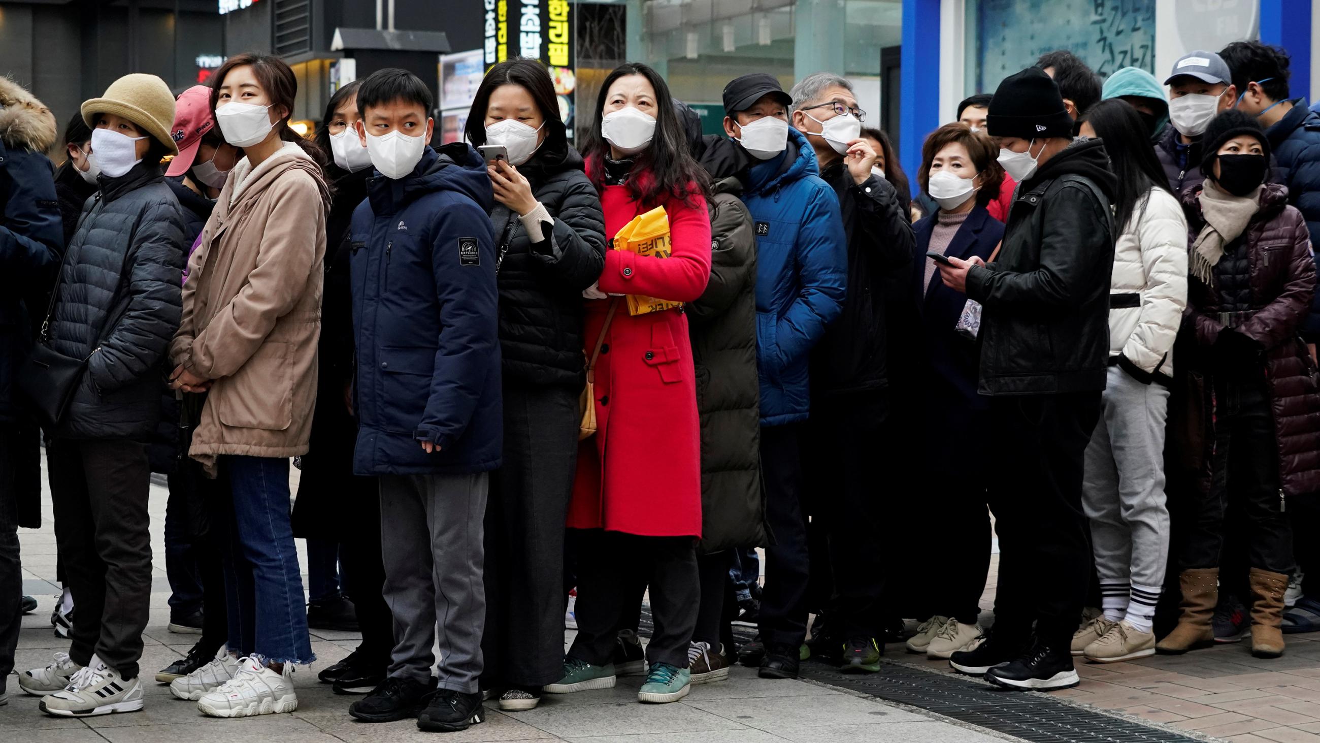 The image shows a long line of people of all ages waiting to purchase masks. They are all already wearing masks. 