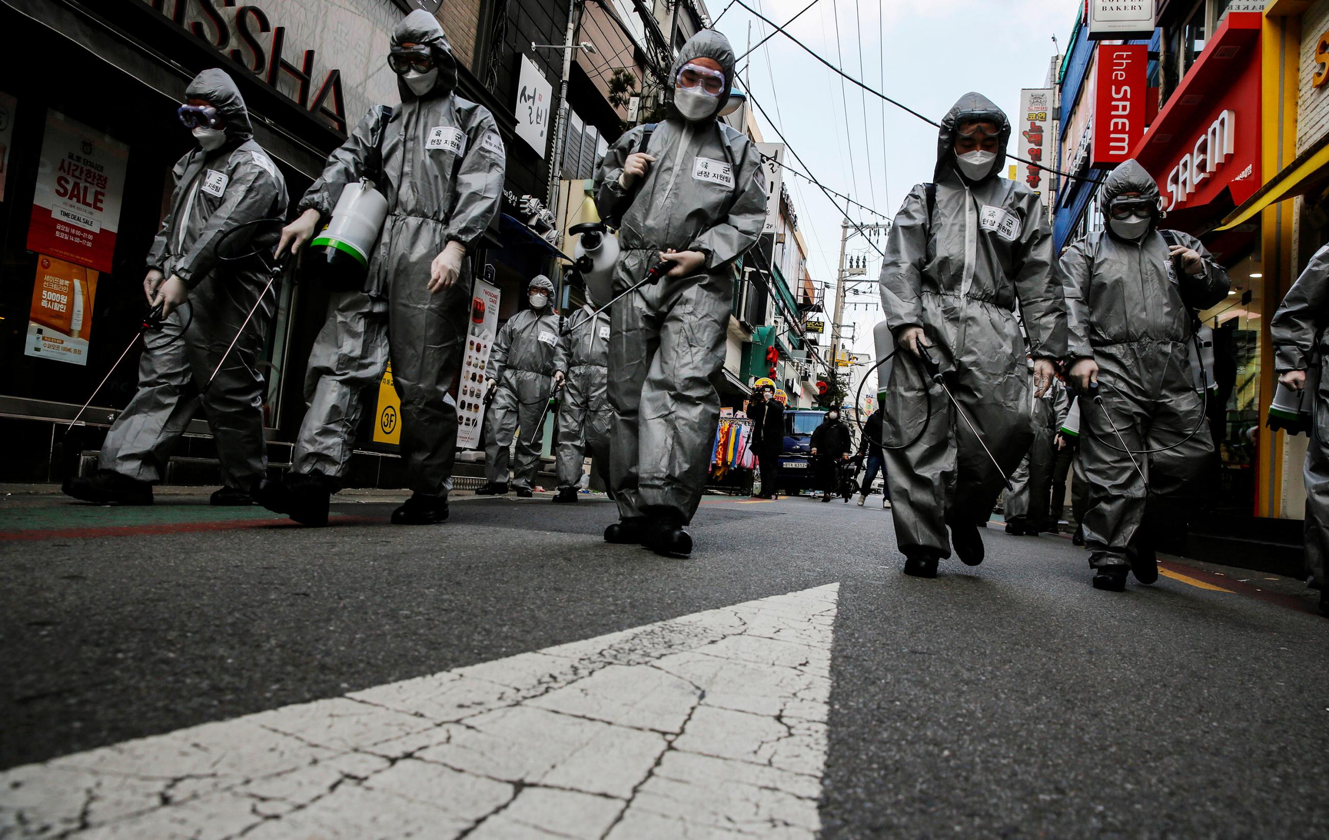 The image is striking showing the soldiers dressed in silver space-suit looking protective gear walking down a more or less abandoned street in what appears to be a normally busy shopping district. 