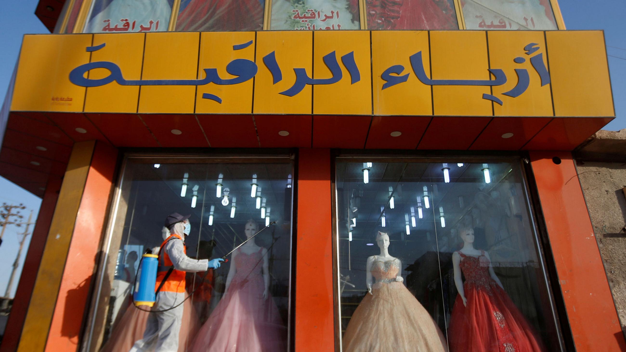 This is an interesting photo showing a colorful dress shop with a worker walking by outside in protective gear spraying disinfectant.