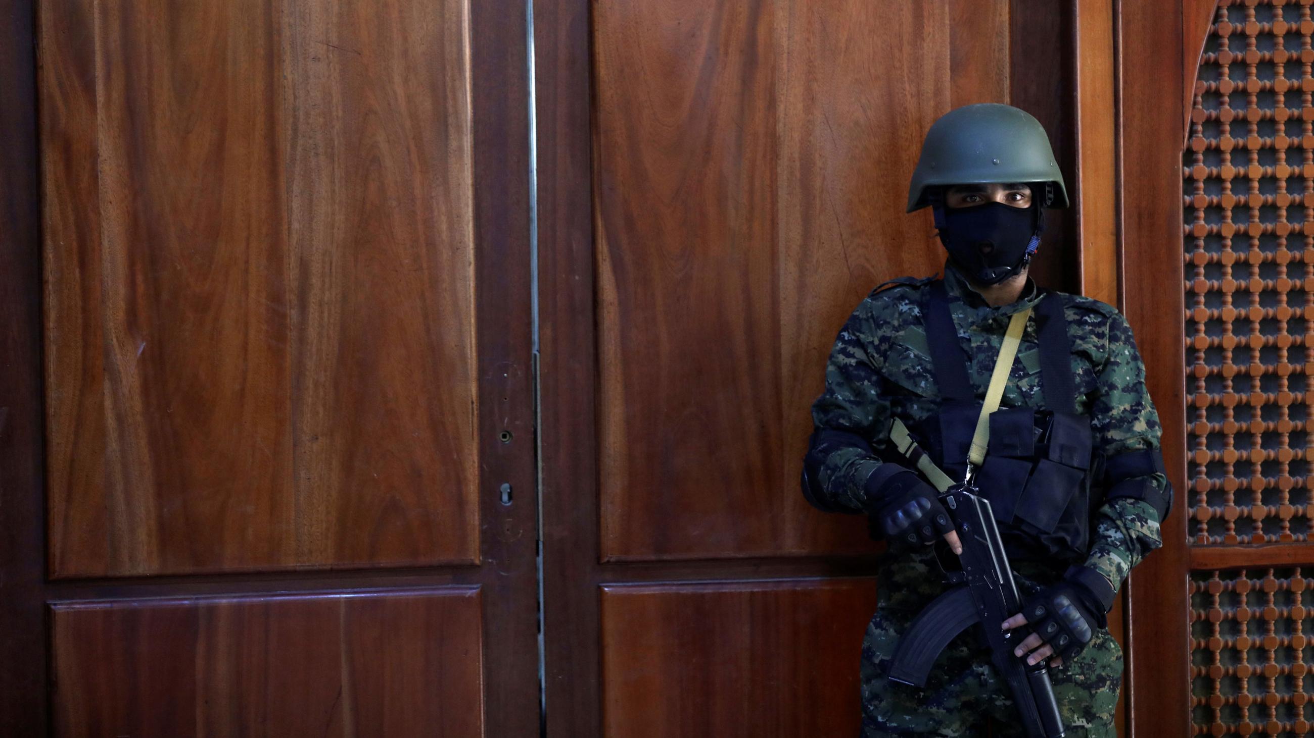 The picture shows a guard with a machine gun standing in front of a wooden door. His mask is black, the door is red.