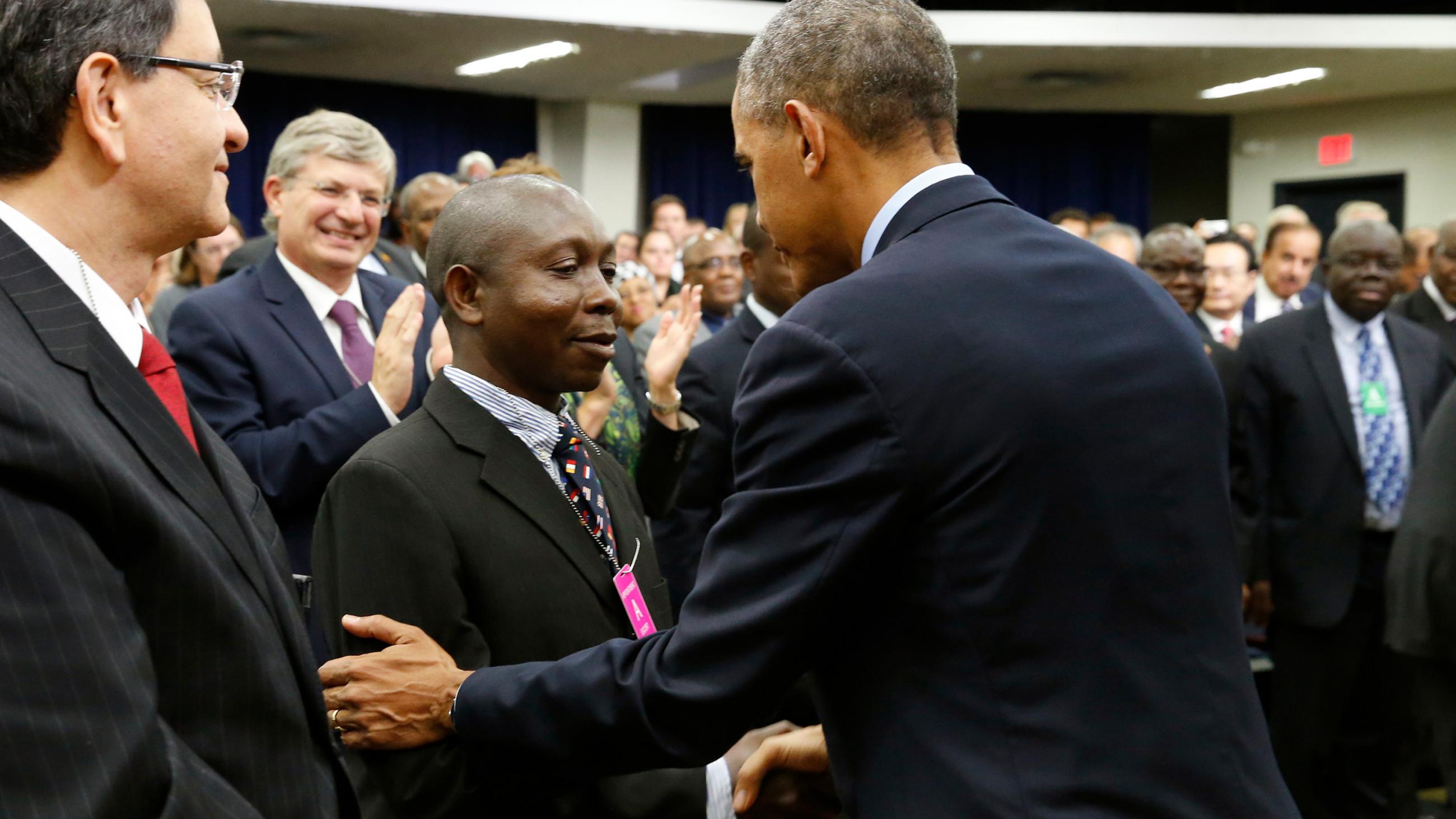 The picture shows the president with his back to the camera shaking hands with a man who is smiling broadly. They are surrounded by other officials and guests. 