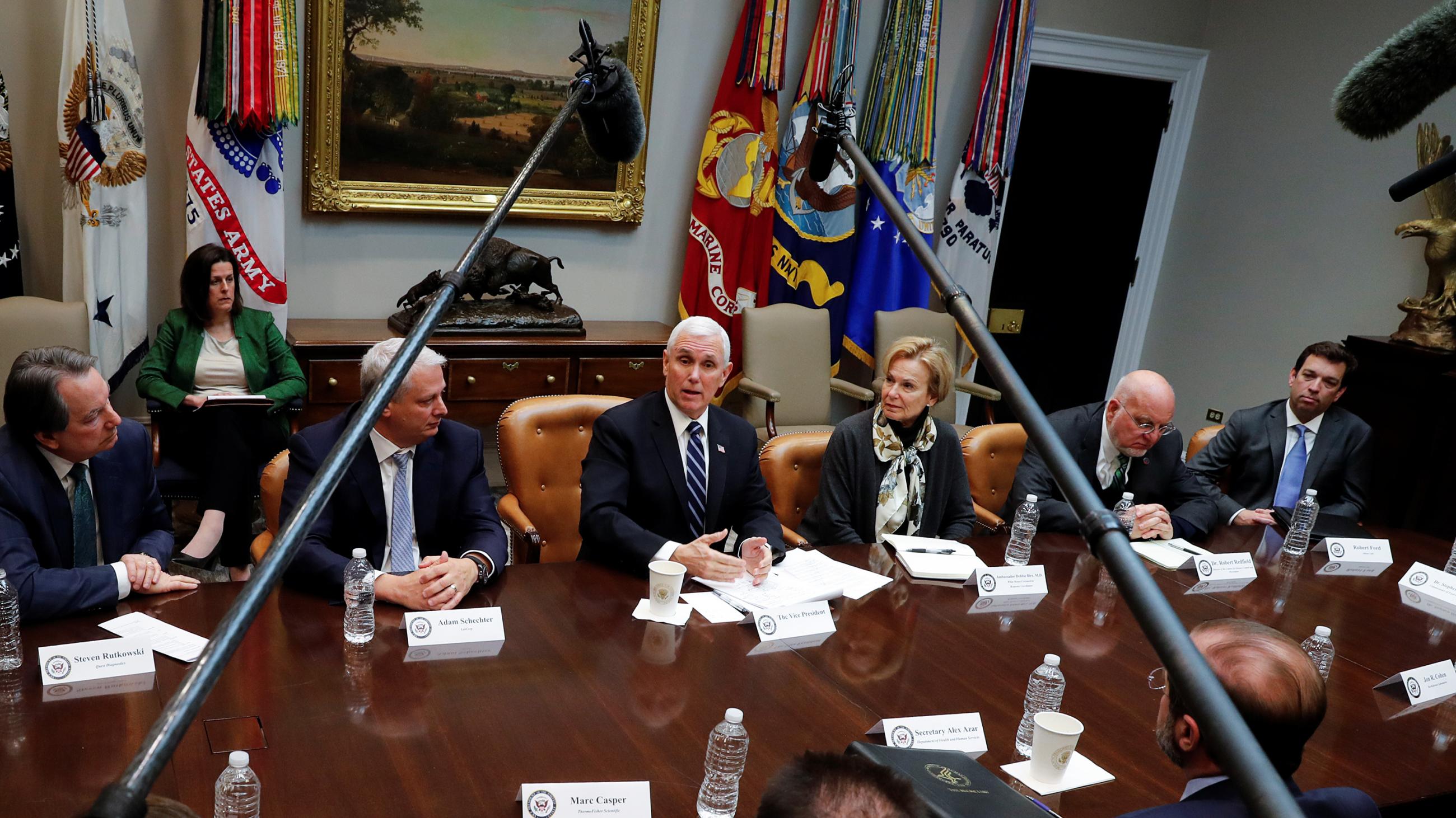 Photo shows the vice president at a large table speaking and surrounded by other people. Two microphone boom poles from opposite ends of the table extend out and converge over the table toward him.