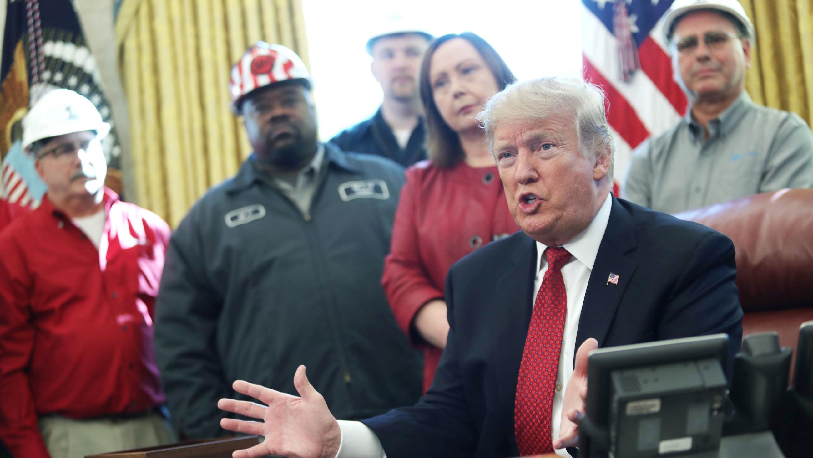 The photo shows the U.S. president at his desk talking and gesturing with his hands as he is surrounded by what appear to be factory workers, wearing brightly colored shirts. Some of them have hard hats. 