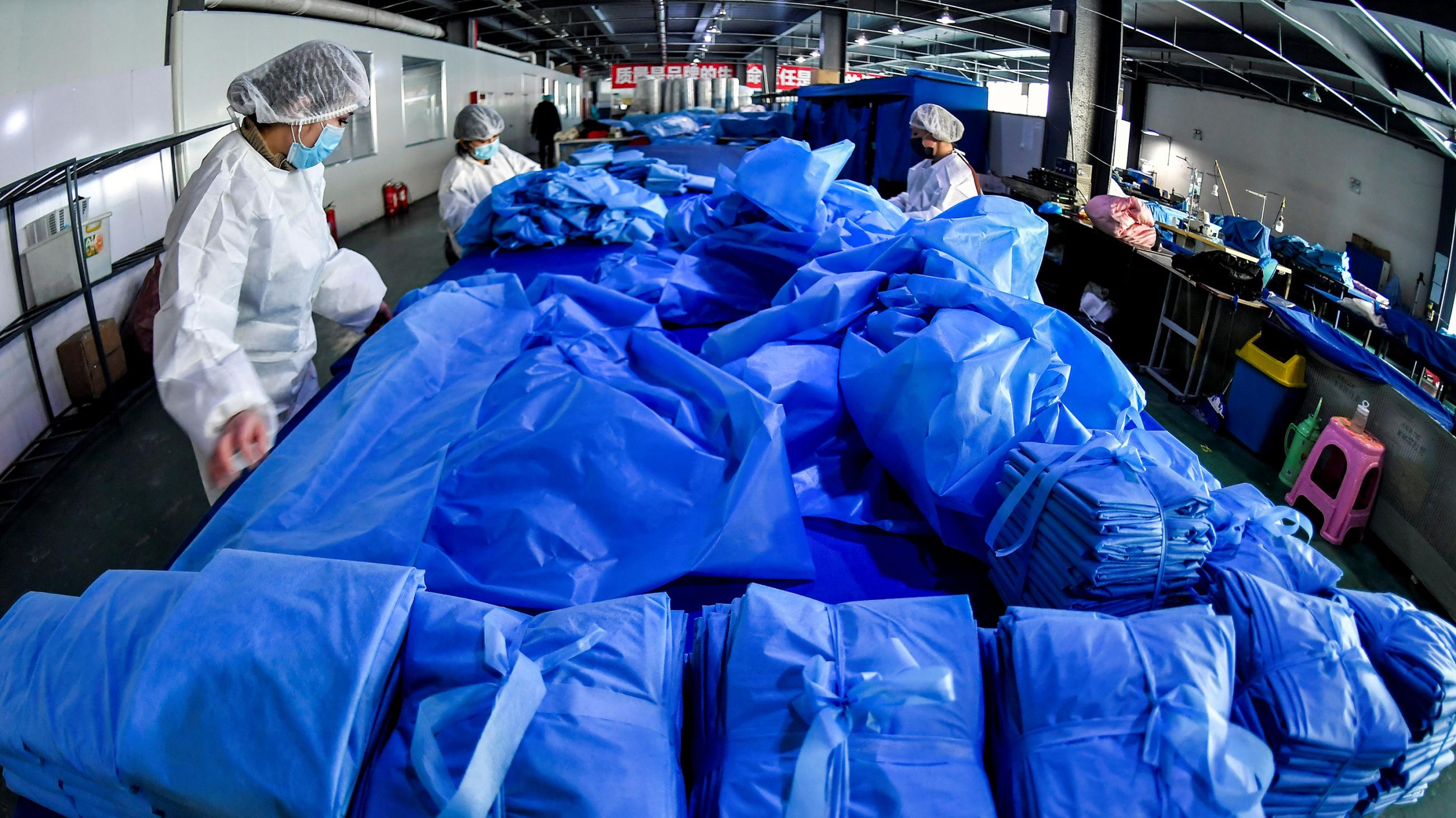 Picture shows workers wearing protective gear standing among piles of folded hospital gowns and other protective equipment.