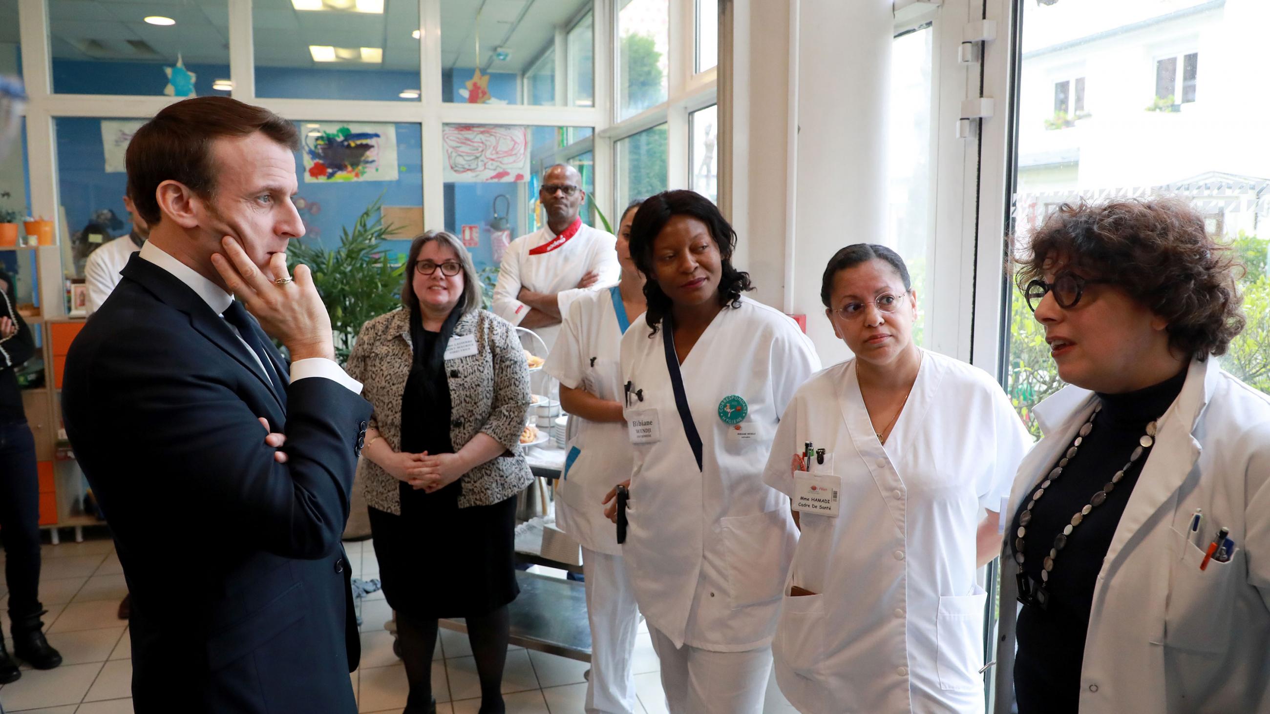 The photo shows about a half dozen people wearing hospital uniforms. One is speaking to Macron, and he is listening intently. 