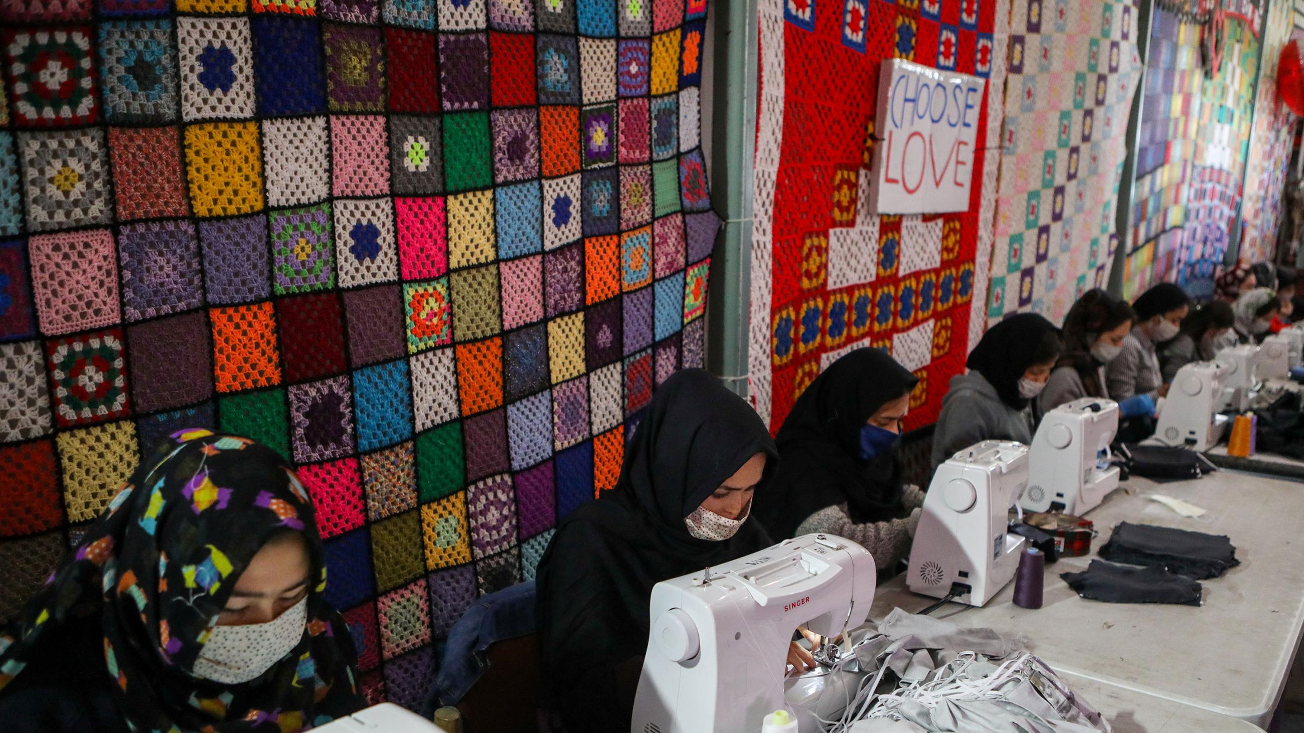 The photo shows a row of women at sewing machines sewing masks with some multicolour quilts in the background. 