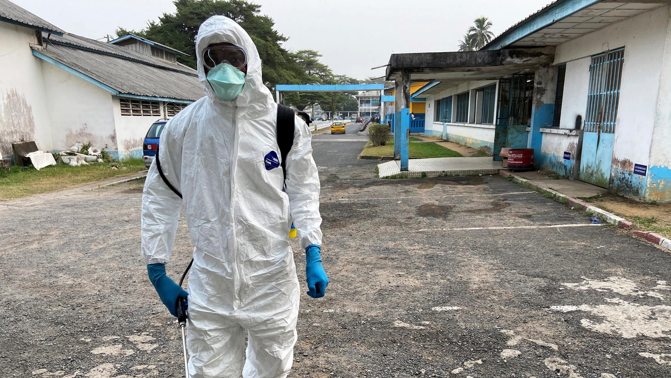 Picture shows the outside of a hospital composed of several low-lying buildings around a courtyard in which a man wearing full protective gear is standing looking at the camera. 