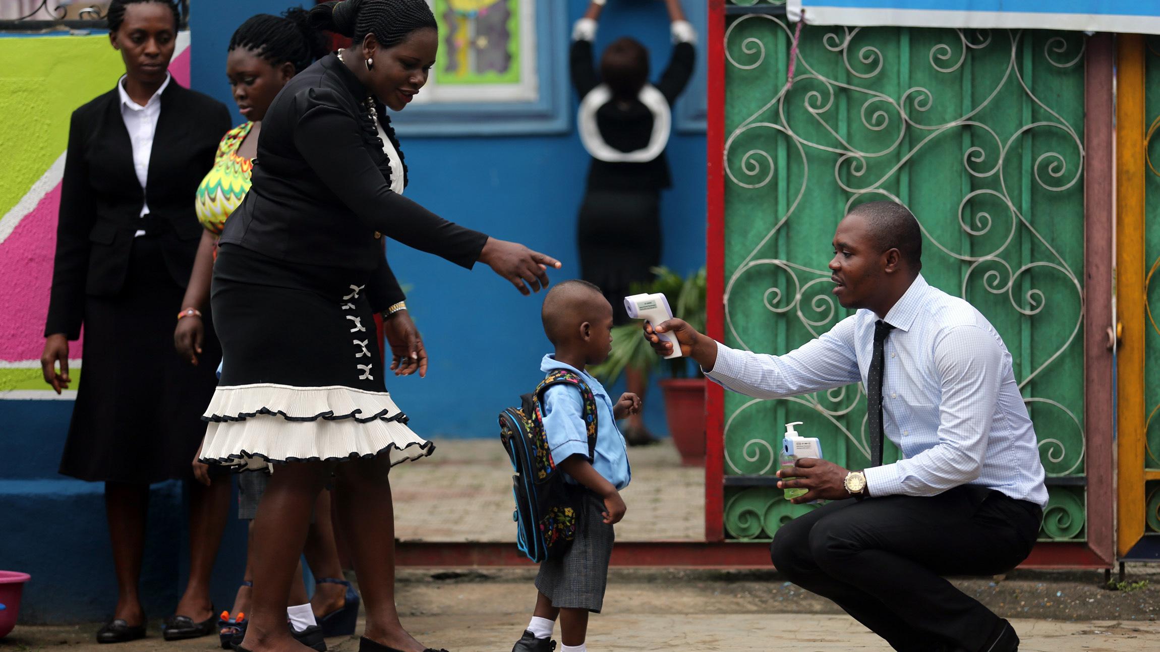 Picture shows a young boy carrying a backpack and wearing a school uniform standing in front of a brightly-colored building. A well-dressed woman directs him while a well-dressed man kneels and points a high-tech thermometer at his head to take his temperature.