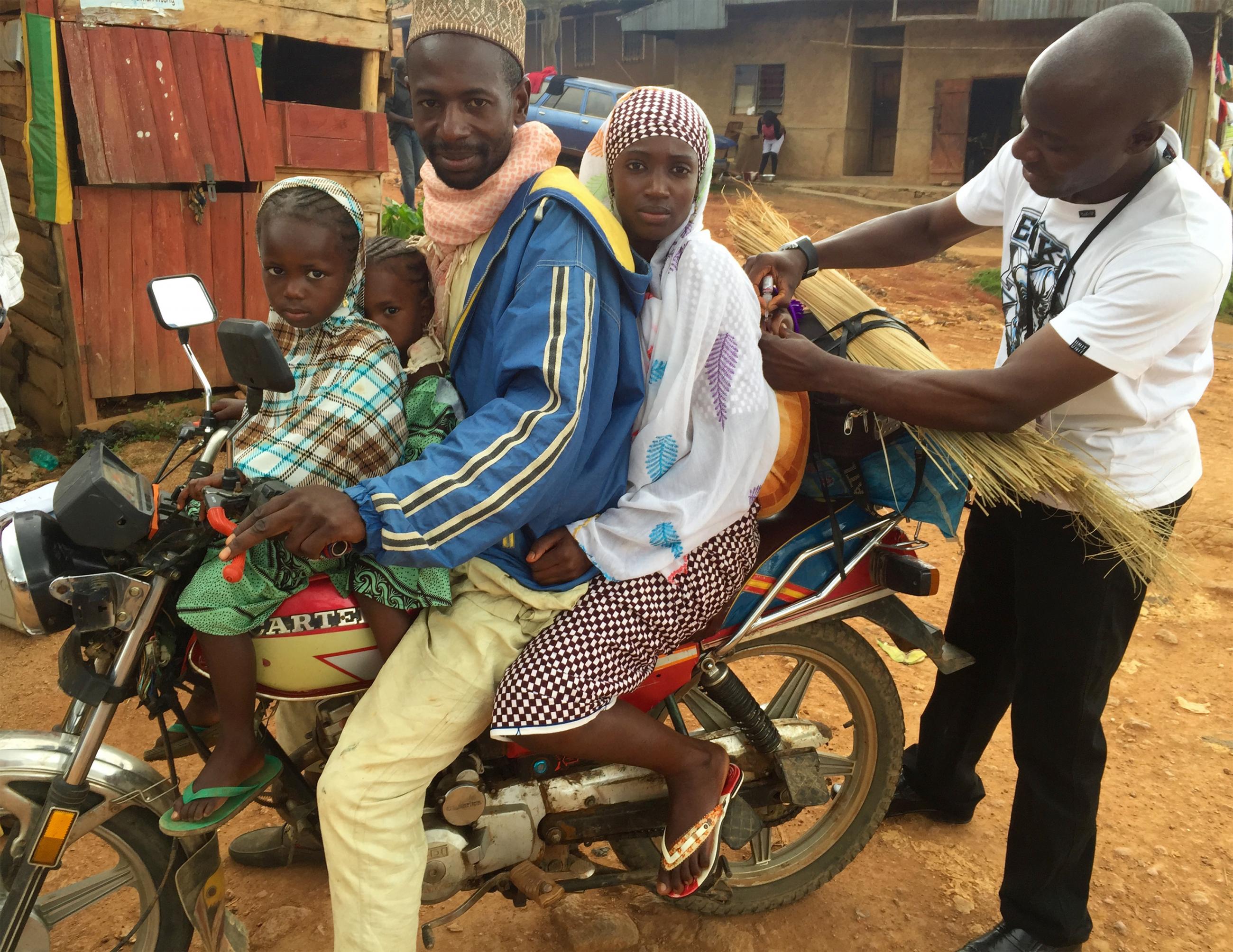 This picture shows a health care worker giving drops of polio vaccine to a child on the back of a motorcycle crowded with both parents and two other small children.