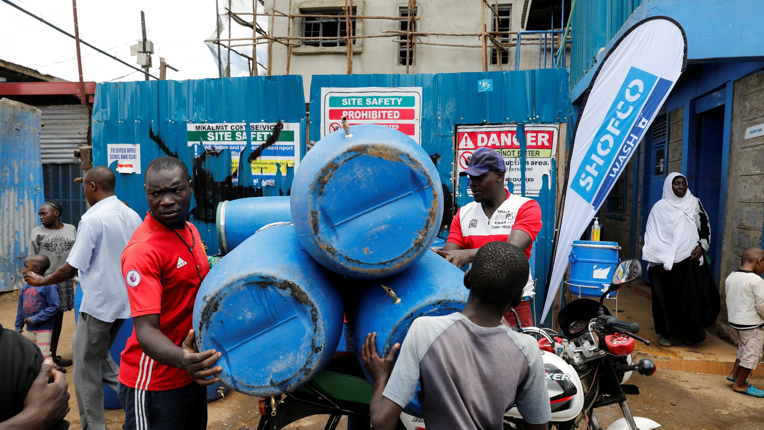The photo shows a crowded street with people loading large blue barrels in the back of a bike. 