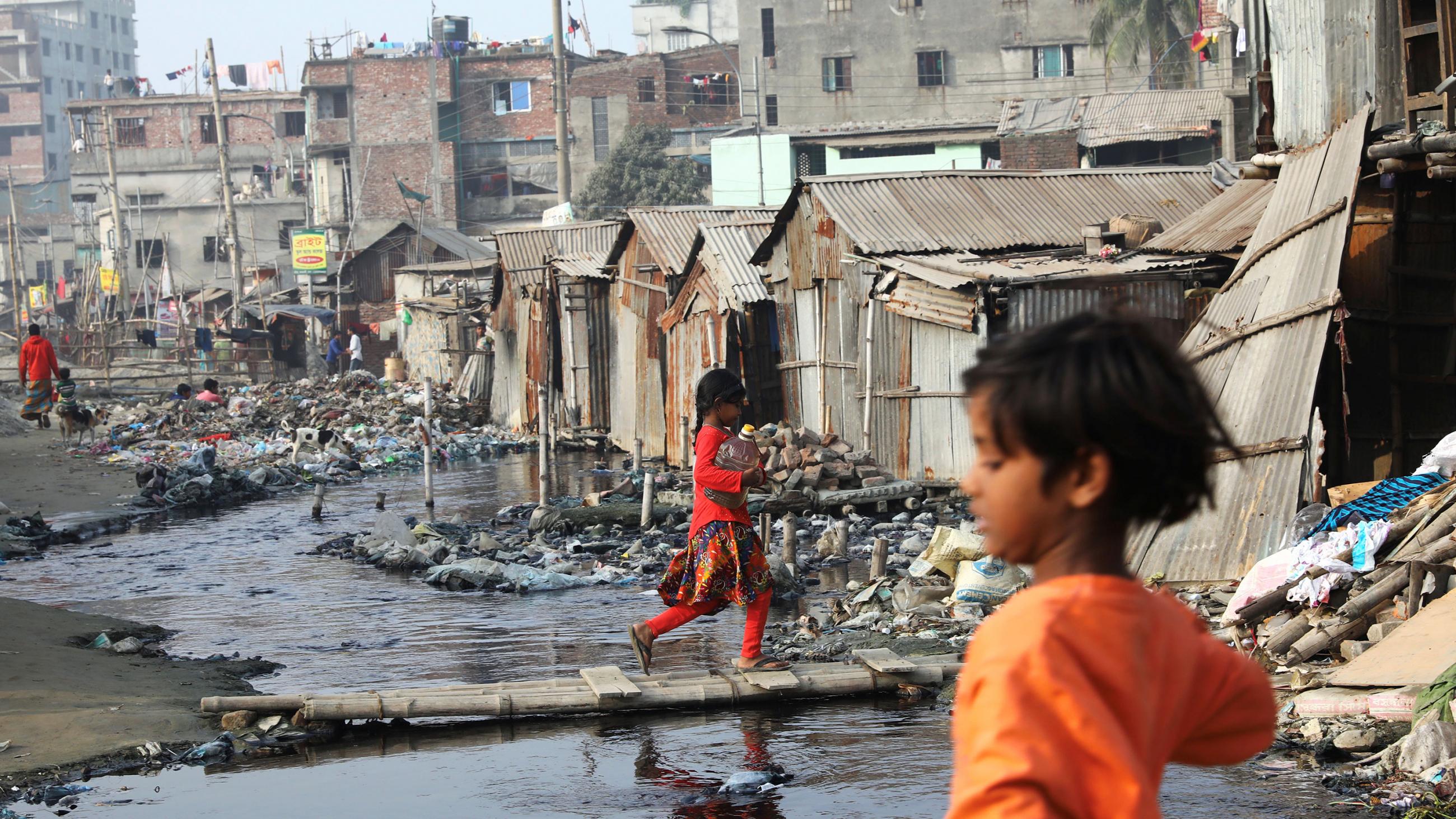 The photo shows a muddy, flooded area in front of tin shacks with a few children walking about. 