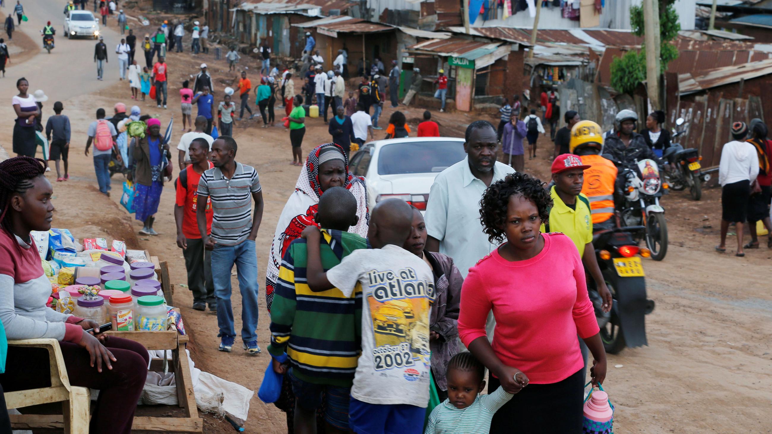 The image shows a long dirt road lined with tin shacks stretching off in the distance. Many people are walking on the road toward the camera and a woman has a table selling goods in the foreground.