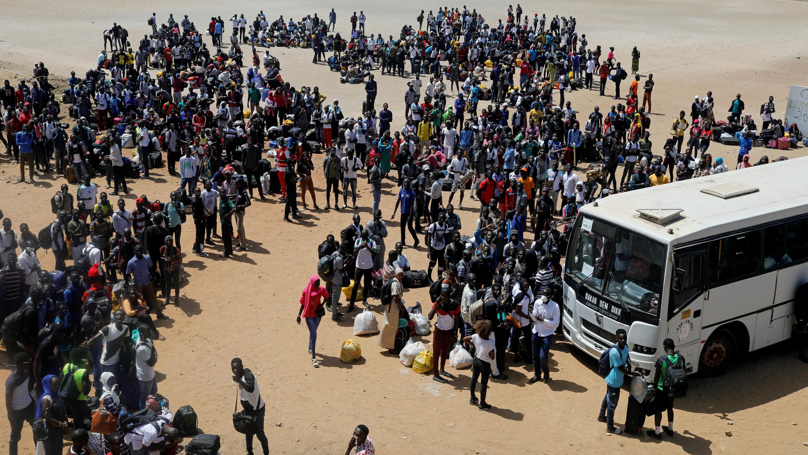 The image is a distance shot of a huge crowd of students with bags in an open area with a single lage bus parked. 