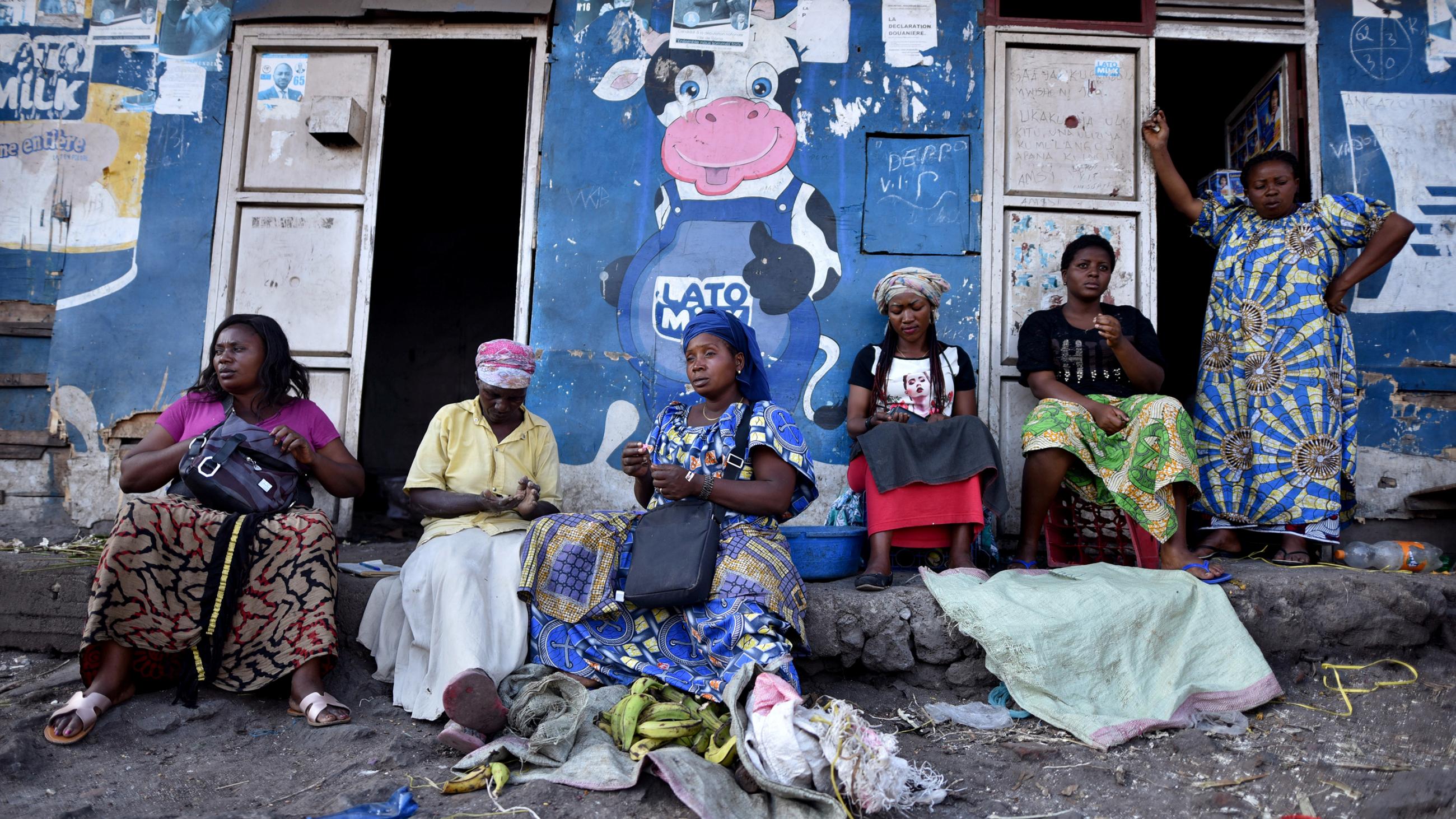 The photo shows a number of women sitting in front of a storefront painted blue and adorned by a large cartoon figure of a cow giving a thumb's up. 