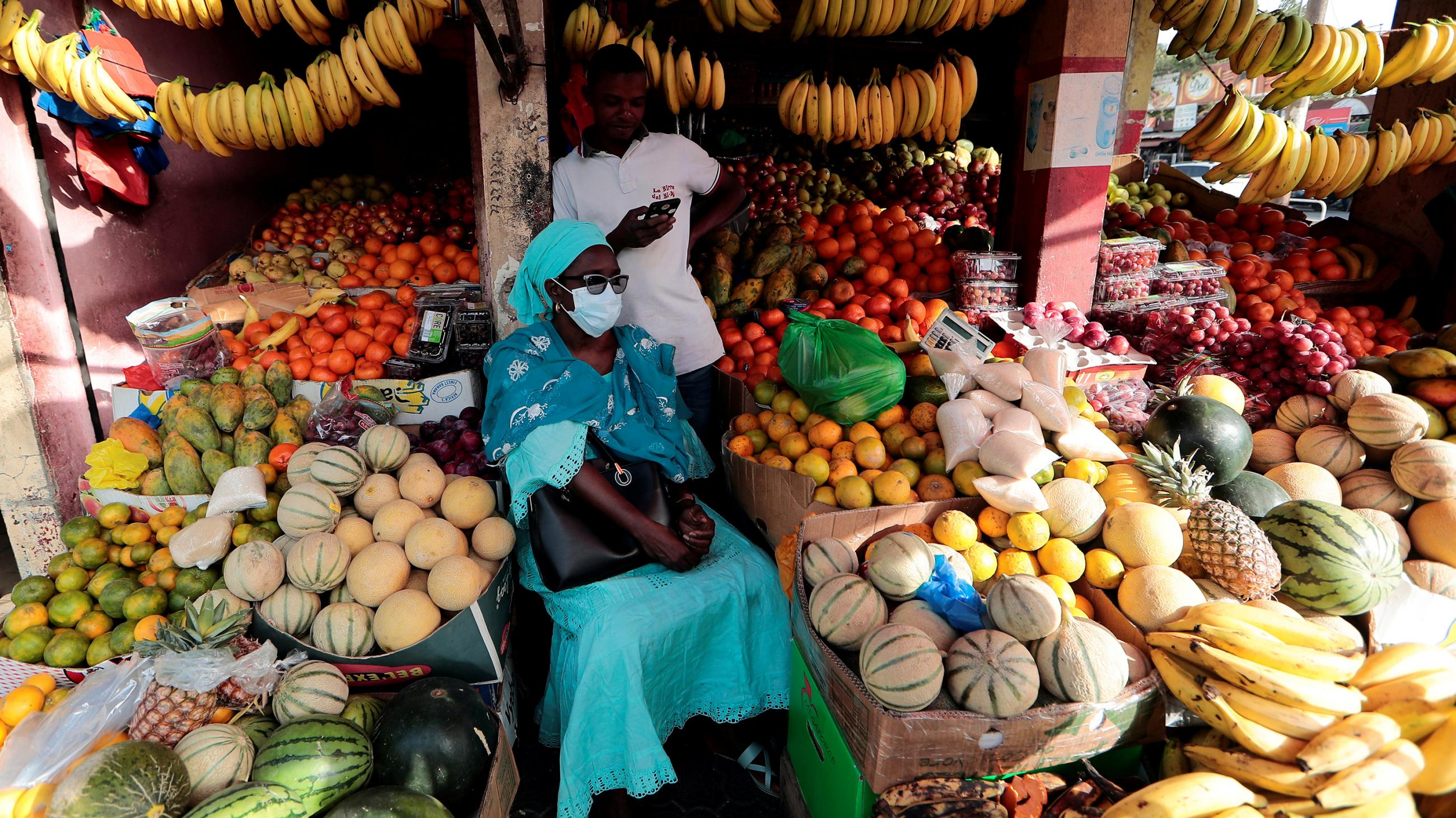 The photo shows a woman wearing a mask sitting amid an abundance of fruit in a tiny stand. 