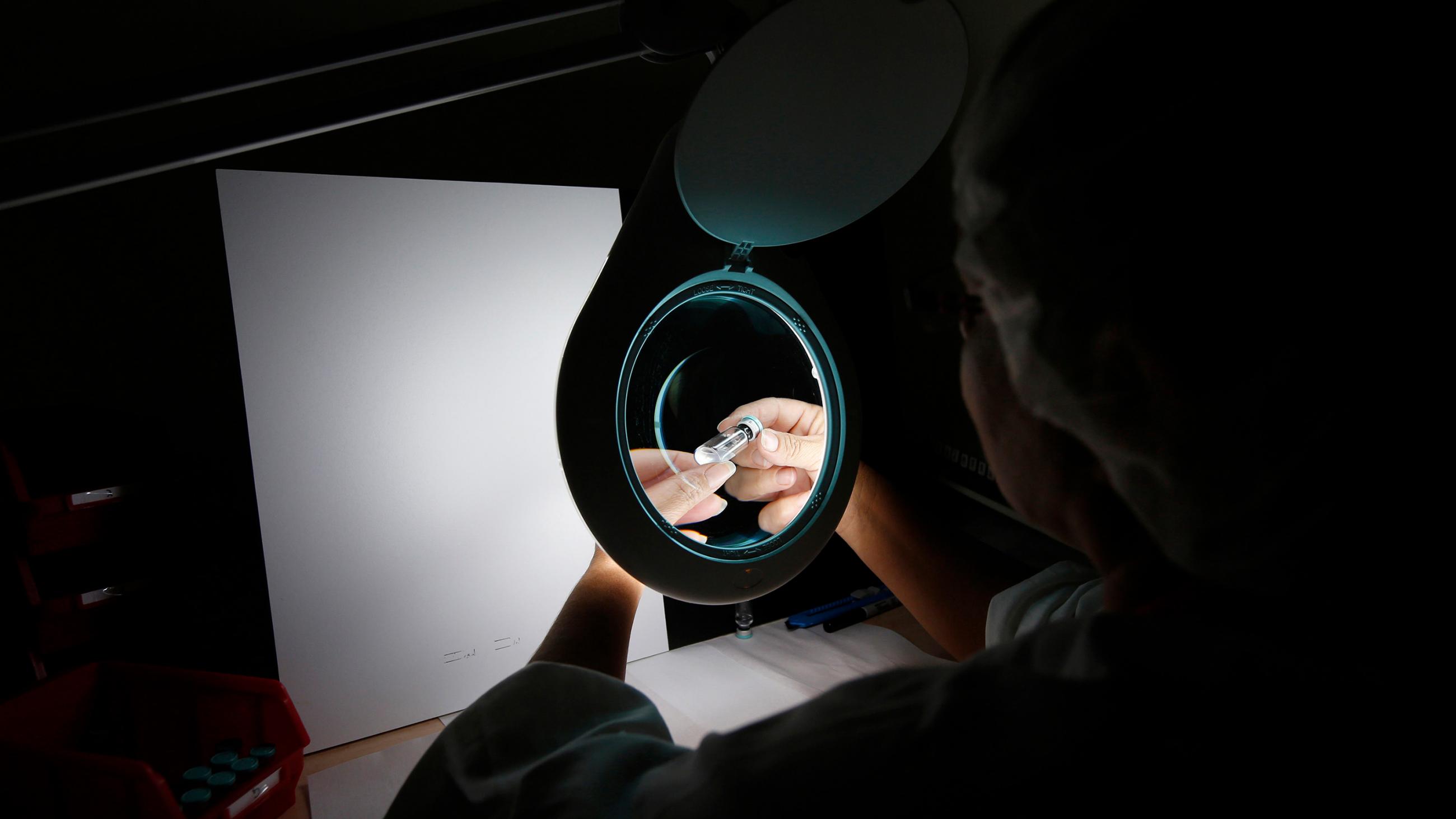 Picture taken shows a laboratory worker at a darkened bench looking at a tiny vial under a lighted magnifying lens. 