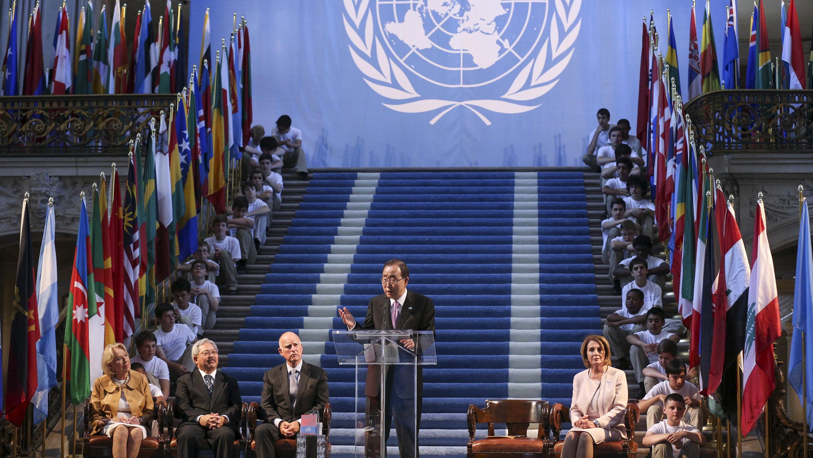 The photo shows the former UN Secretary-General at a podium on an elaborately designed stage with steps leading up and children dressed in white shirts and tan slacks on either sides of the stairs. 