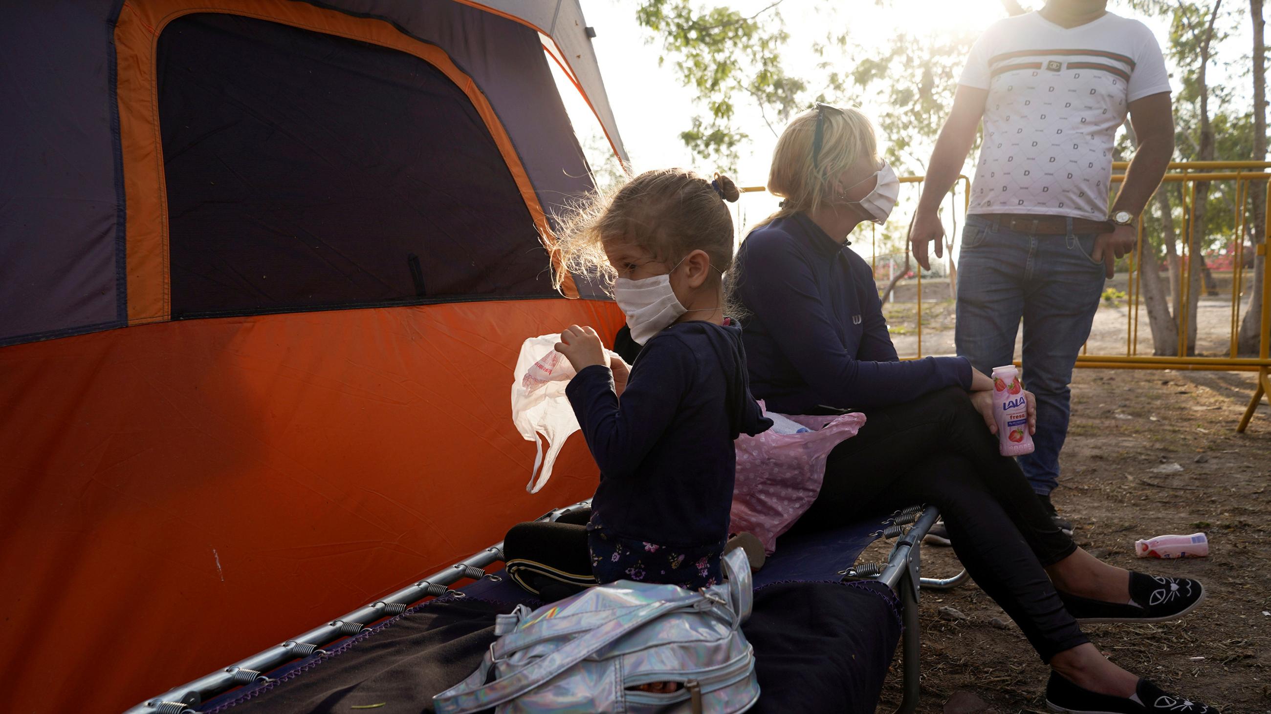 The photo shows the mother and daughter outside a tent in the morning hours with the rising sun brightly illuminating the top of the frame. Mother holds a strawberry drink and talks to a man whose head is out of the frame. Daughter faces the other way and plays with a plastic shopping bag. 