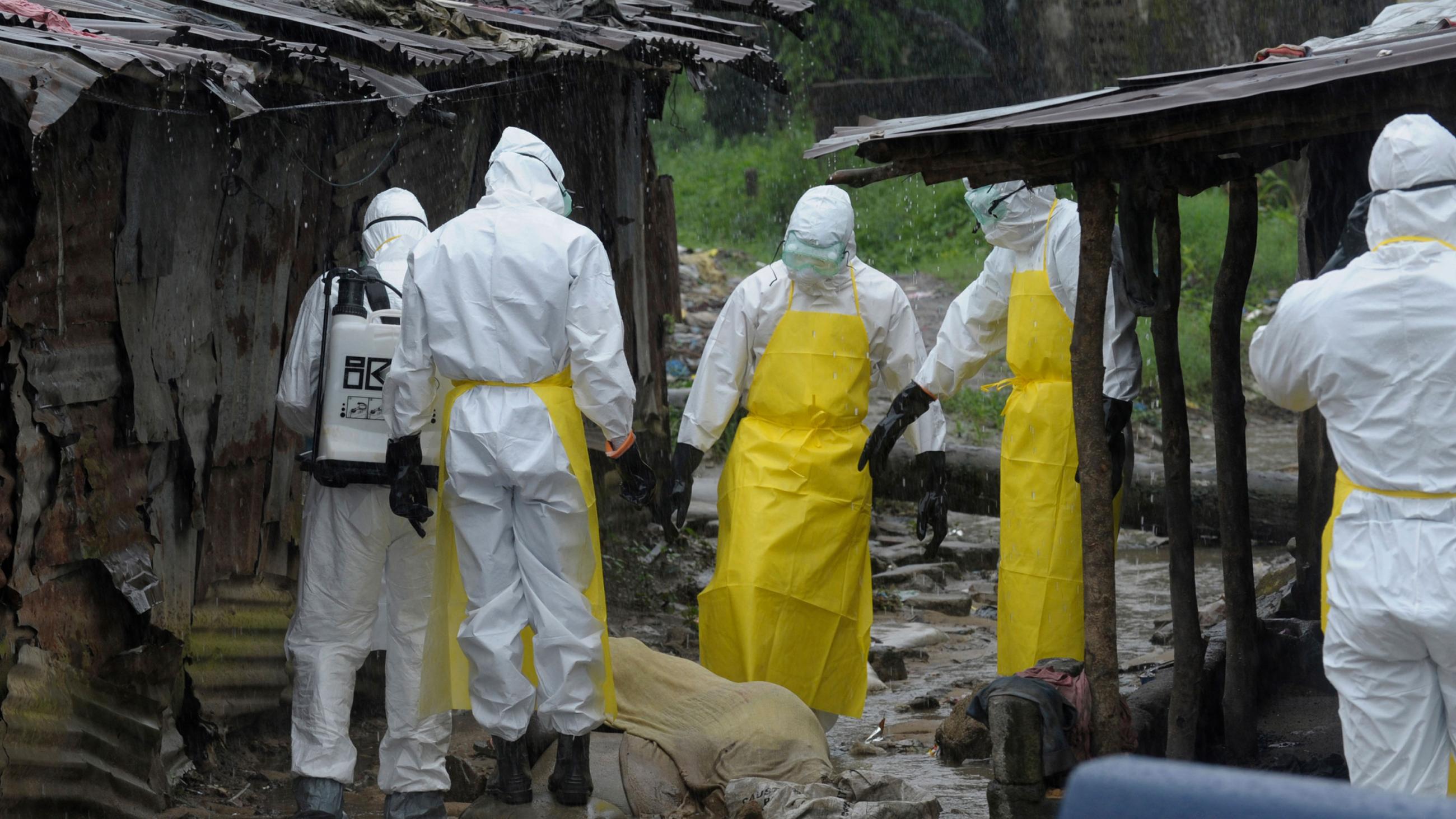 The photo shows half a dozen workers in full body protective suits standing in large mud puddles in the driving rain beside a body draped by a thin muslin cloth in between some ramshackle shacks. Some are pointing, and they appear to be discussing how to pick up the deceased victim. 