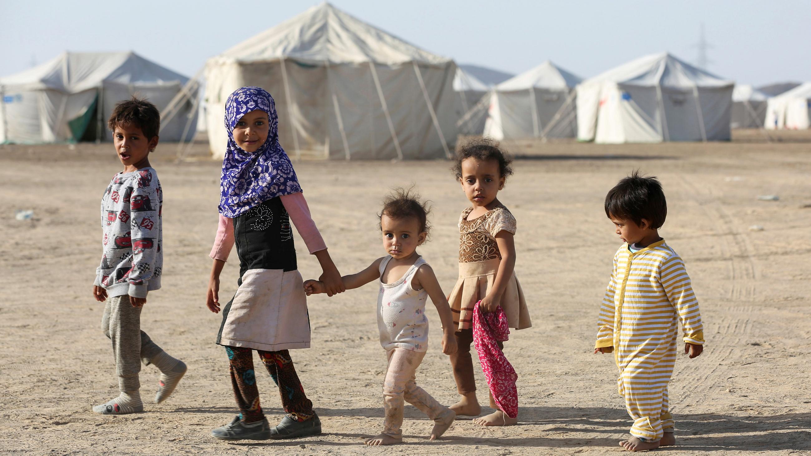  Picture shows a line of five children walking together in front of a camp filled with white tents and looking at the camera. All are very young, two who appear under eight years old, and three who appear under five. 