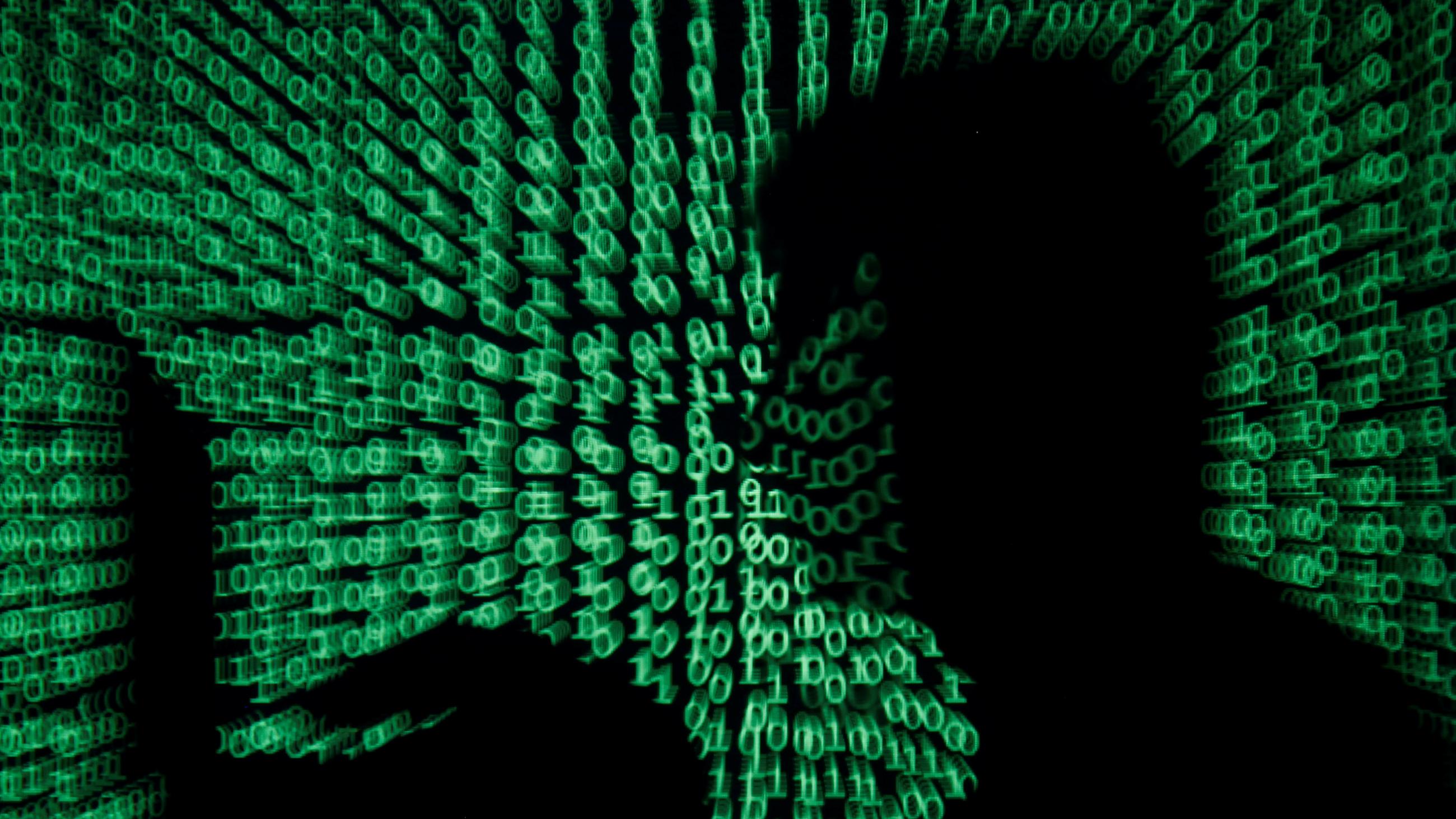 the image shows the silhouette of a man on a laptop with green ones and zeroes, reminiscent of the film "The Matrix" projected over the image. 