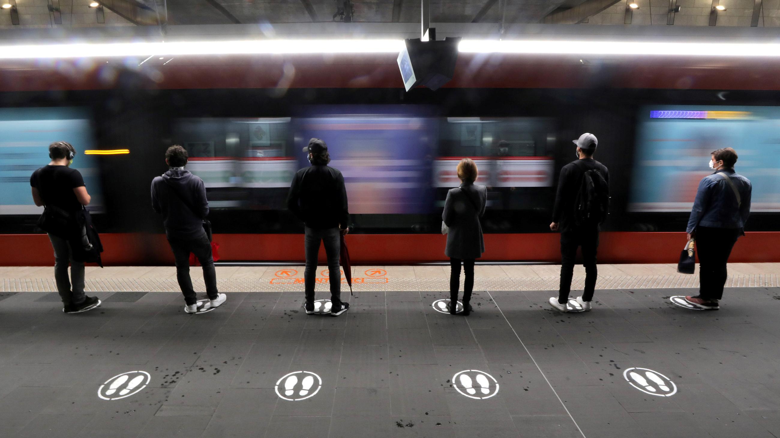 The photo shows people standing on paintef signs of feet on the platform with their backs to the camera as a train rushes by. 