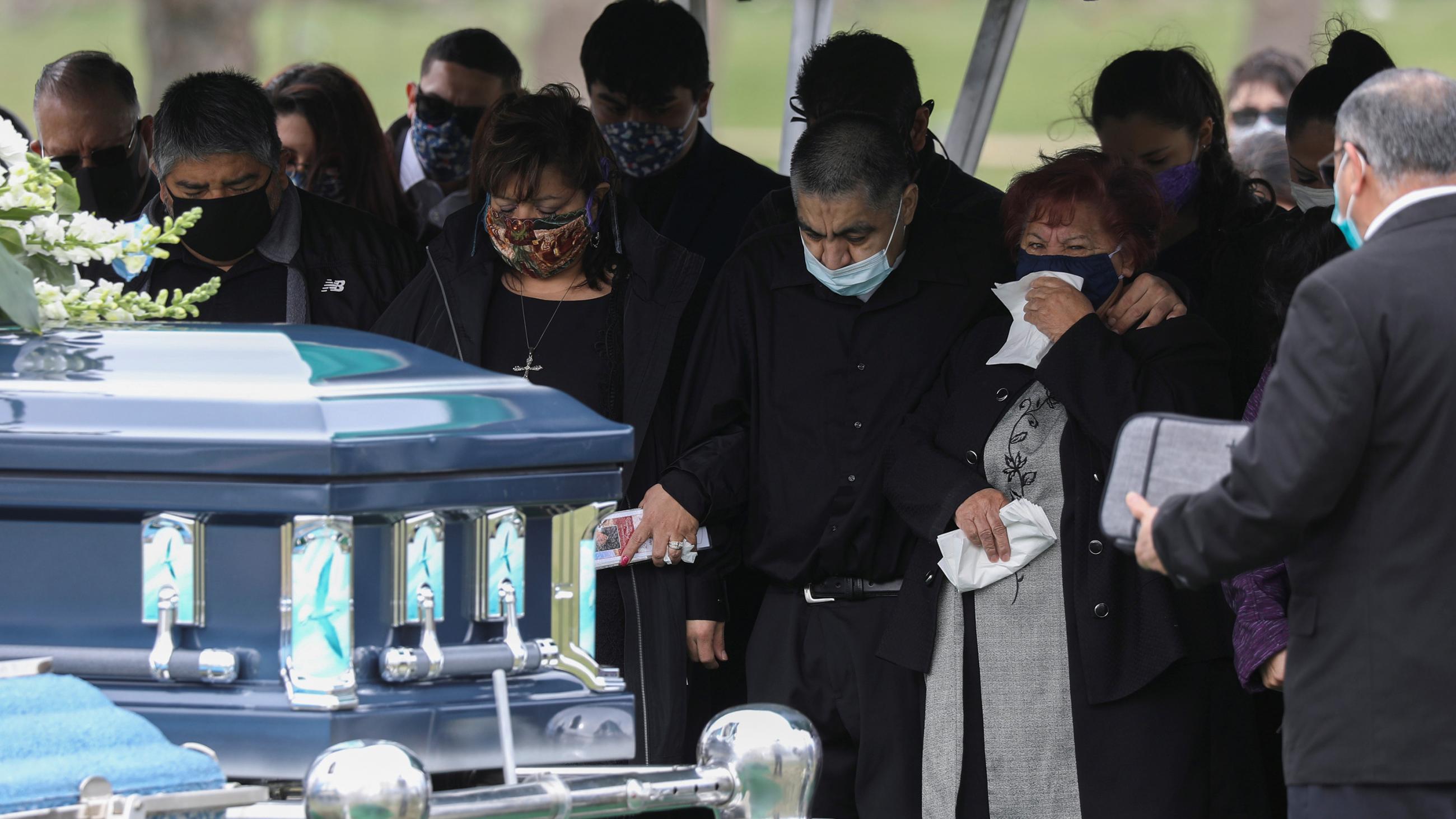 The photo shows a funeral with numerous people weeping for their lost loved one. A casket stands close to the camera, ready to be lowered into the ground. 