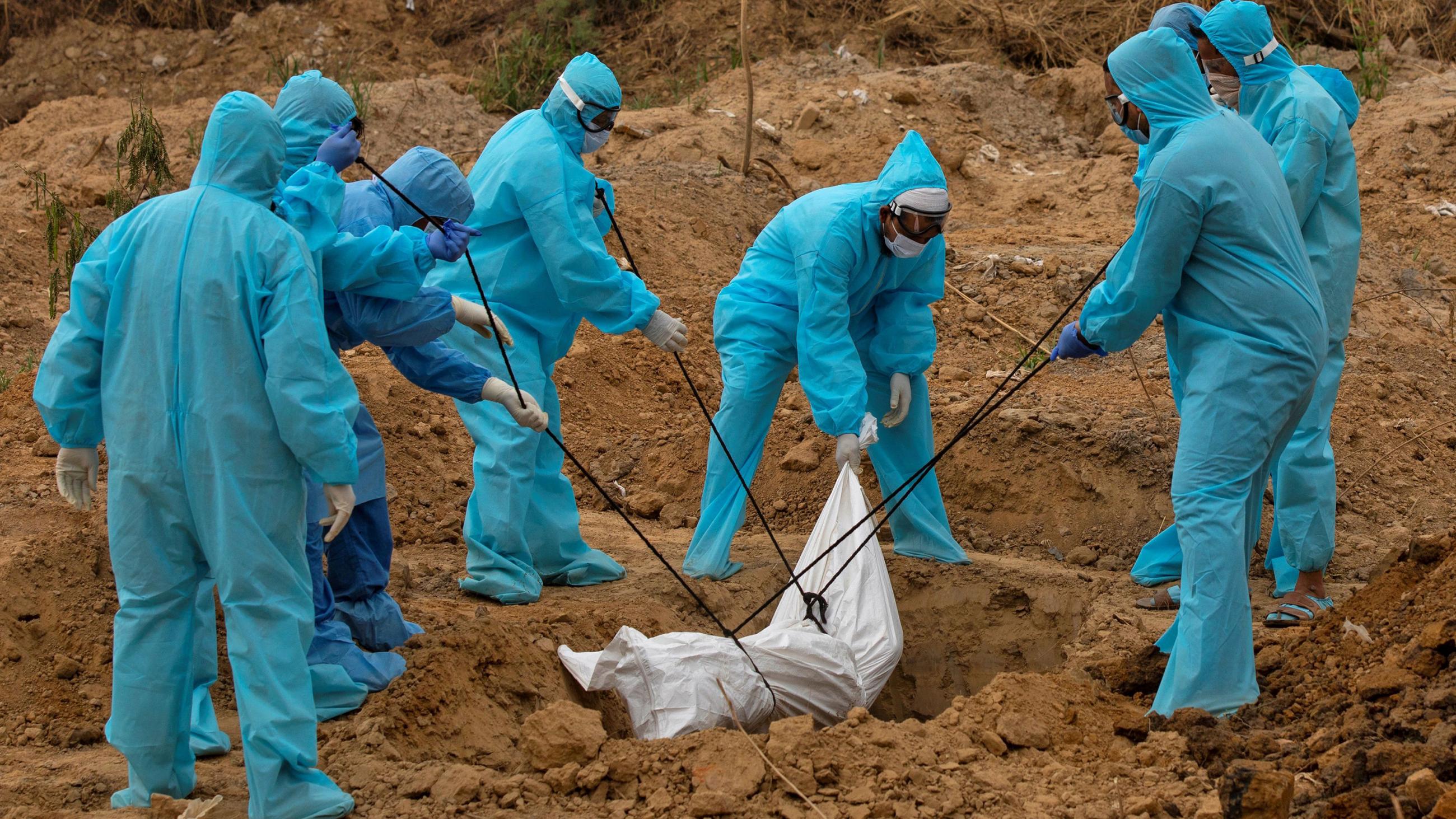 The photo shows a number of people in bright blue protective suits lowering a white sheet-wrapped body into the ground. 