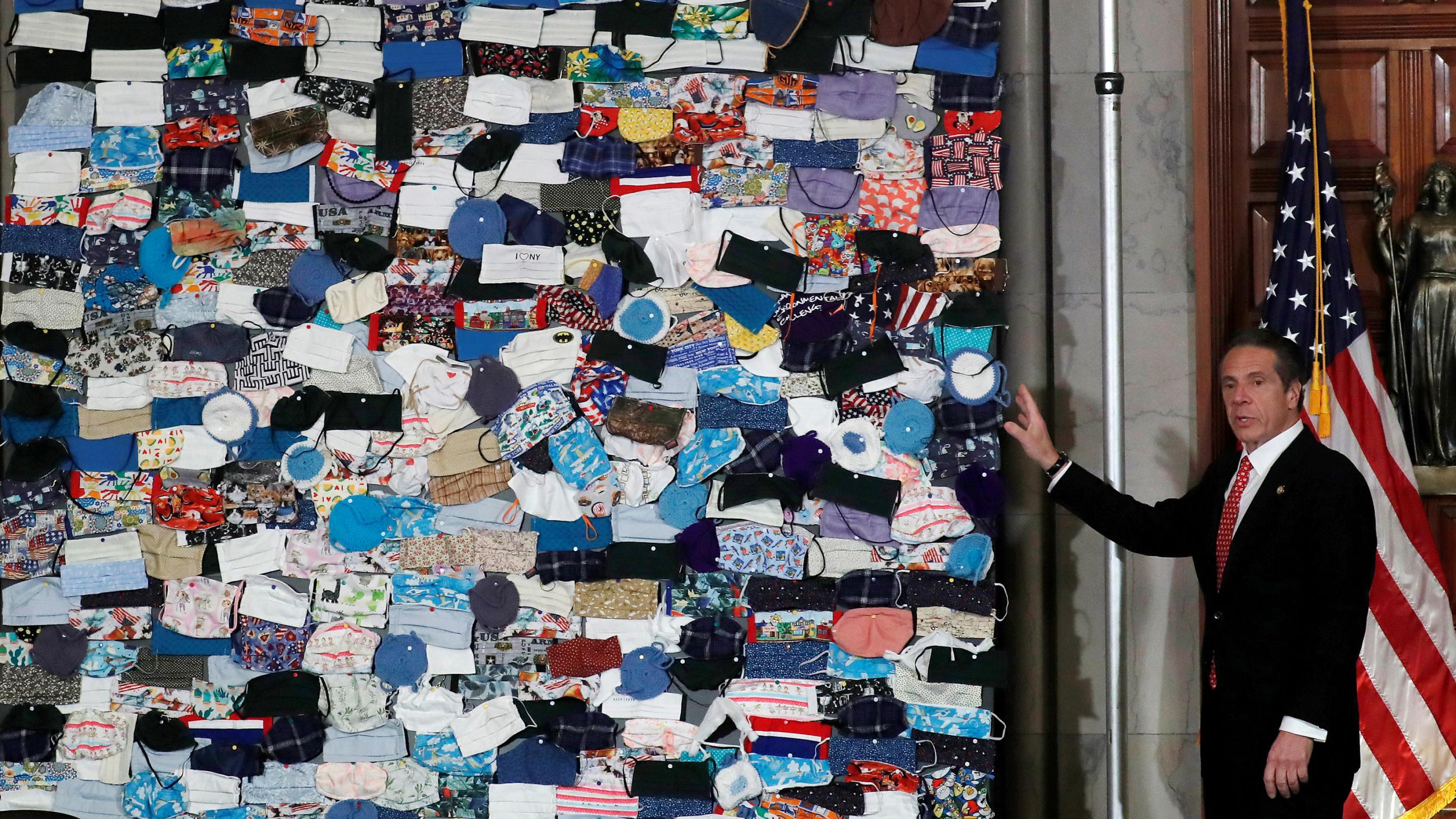 The photo shows the governor in front of a large quilted patchwork of masks. 