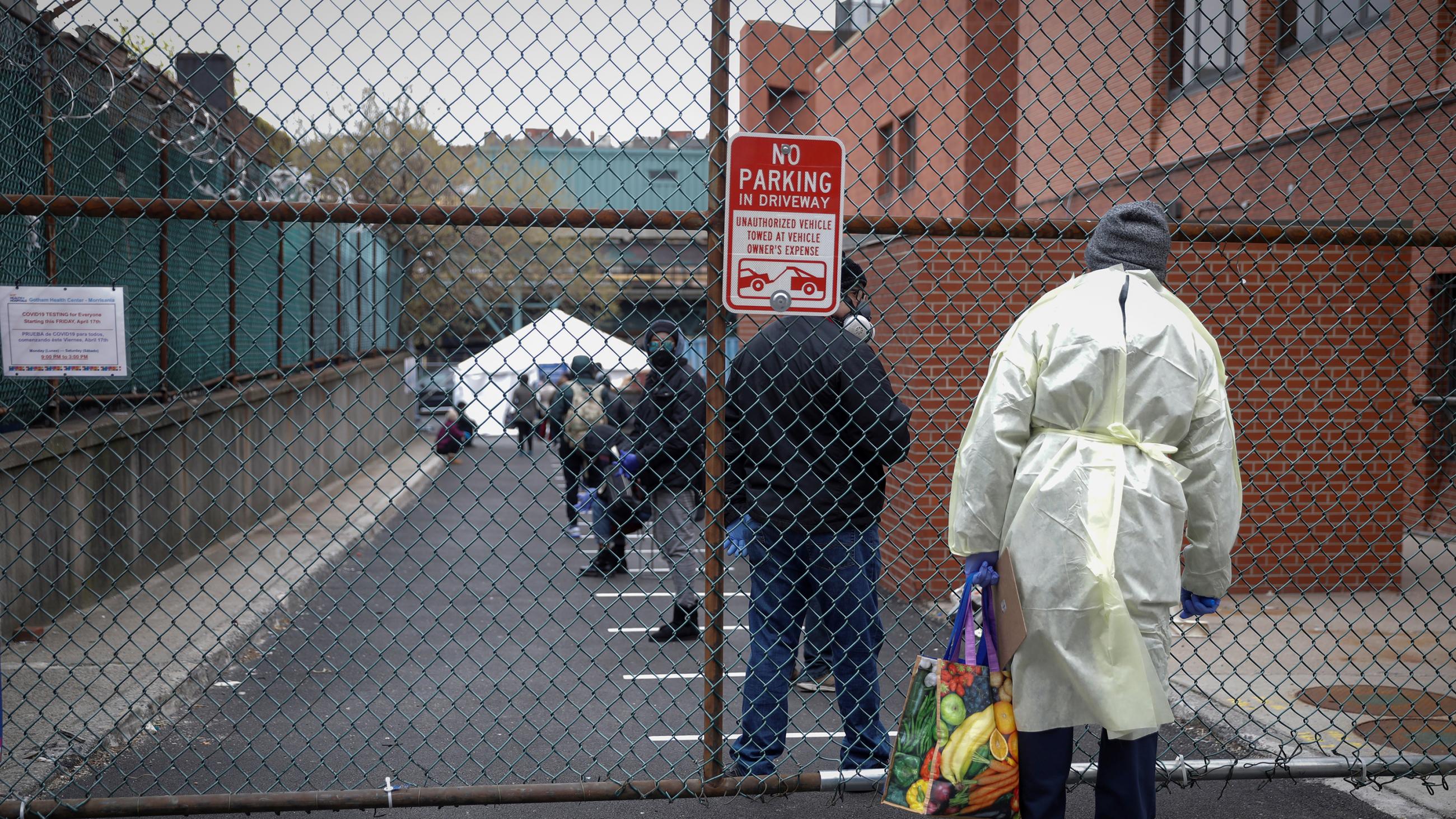 The photo shows a health worker standing behind a fence looking in on an enclosed parking lot where people are lined up awaiting testing. 