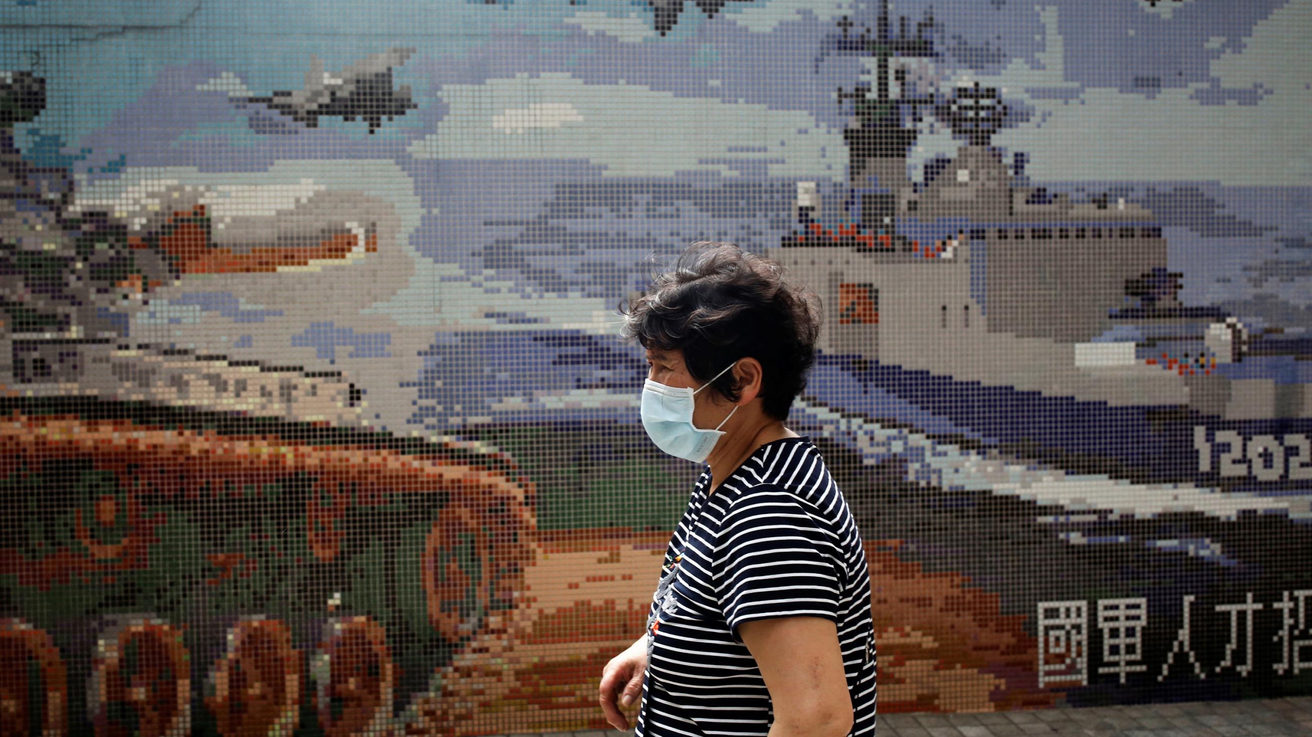 The photo shows a woman in a mask walking past a stylized poster showing war tanks, ships and planes. 