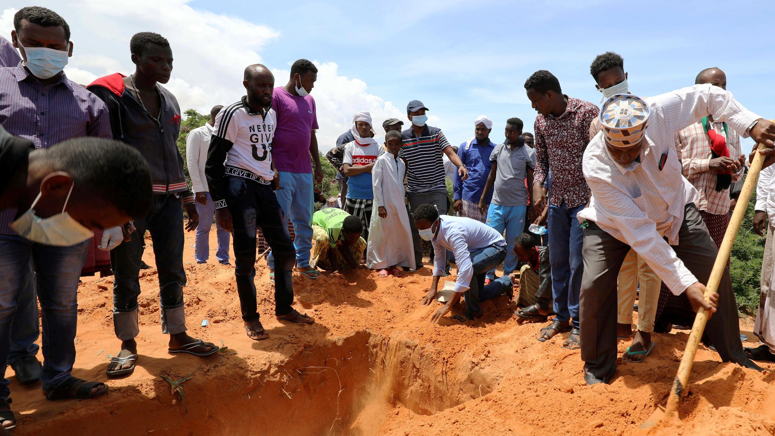The photo shows a crowd gathered around a grave while one person shovels dirt in the hole. 