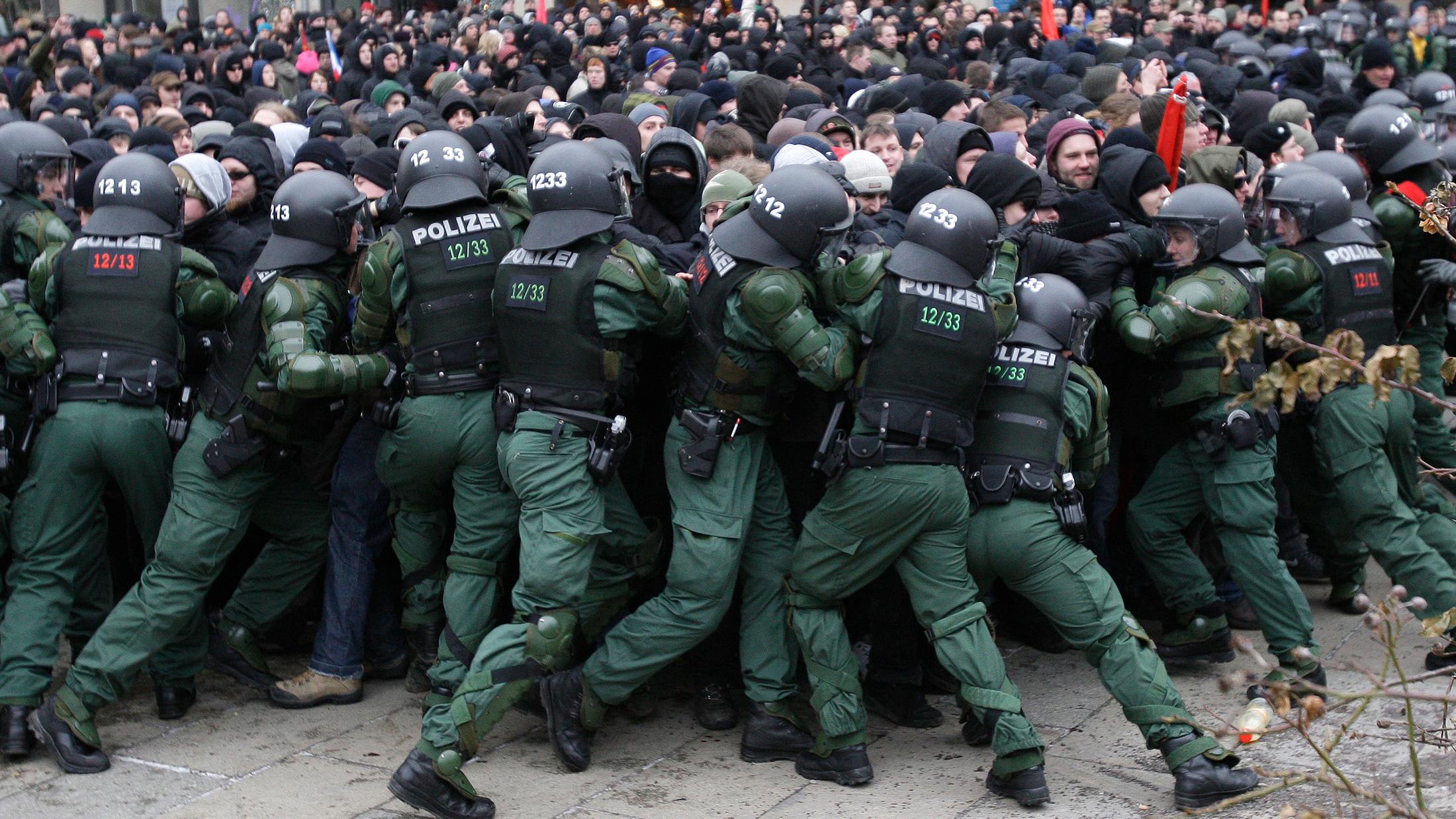 The photo shows a line of uniformed riot gear officers pushing hard against a scrum of protesters.