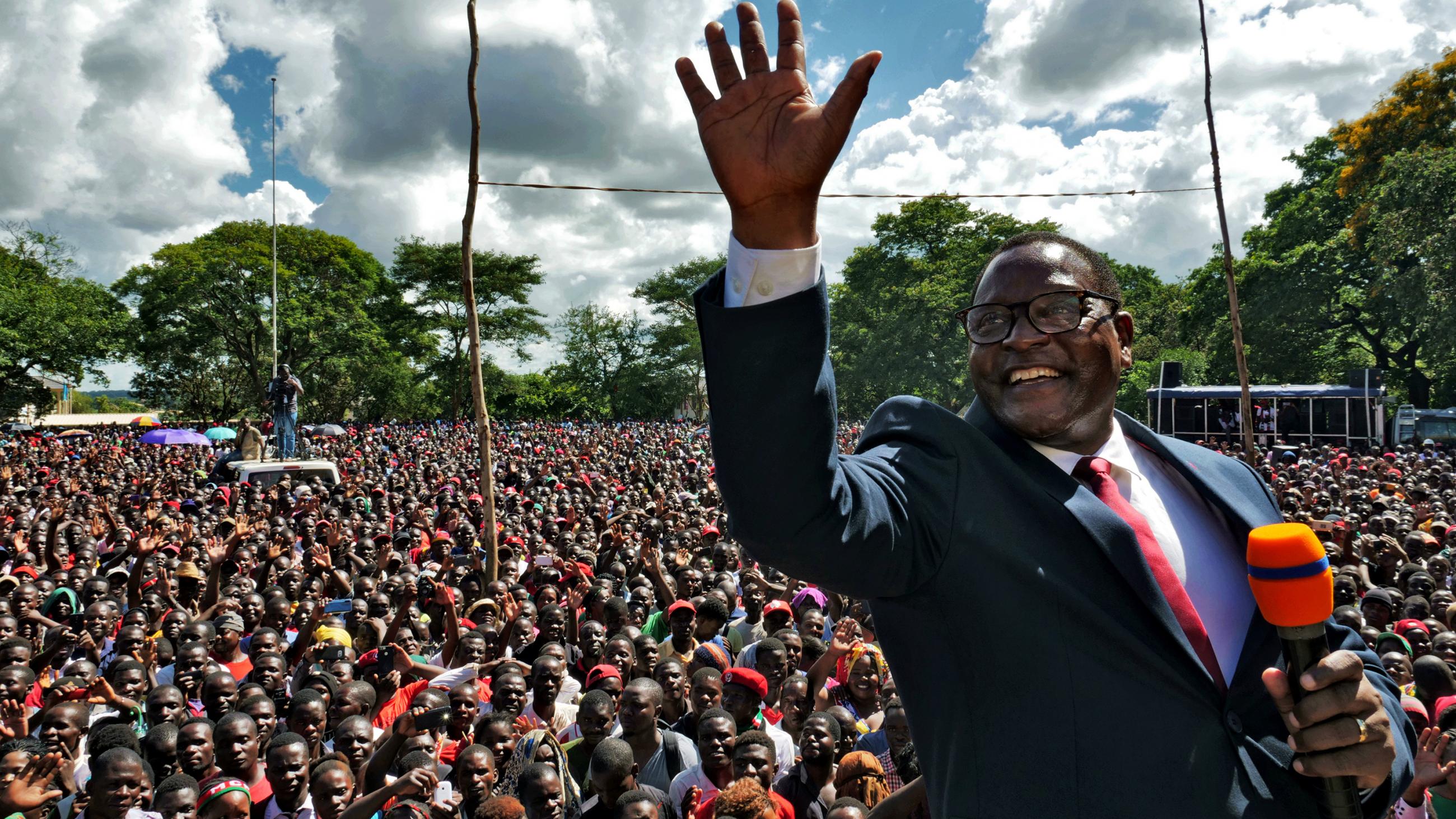 The photo shows a smiling Chakwera, microphone in hand, standing atop a stage waving and smiling. A massive crowd can be seen in the background. 