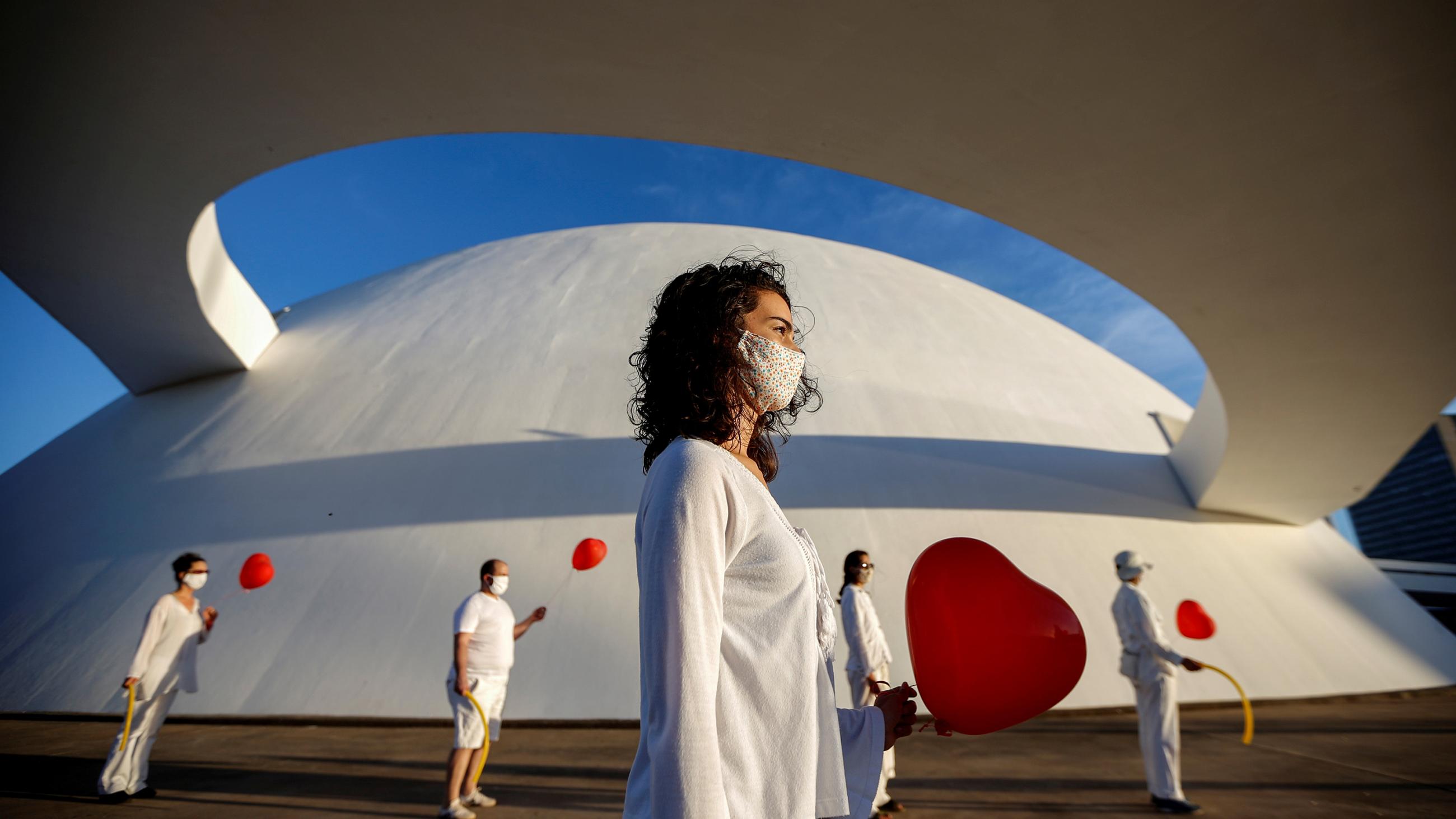 The photo shows a performance art piece with a woman wearing white in the foreground and four people wearing white and holding red balloons in the background. 