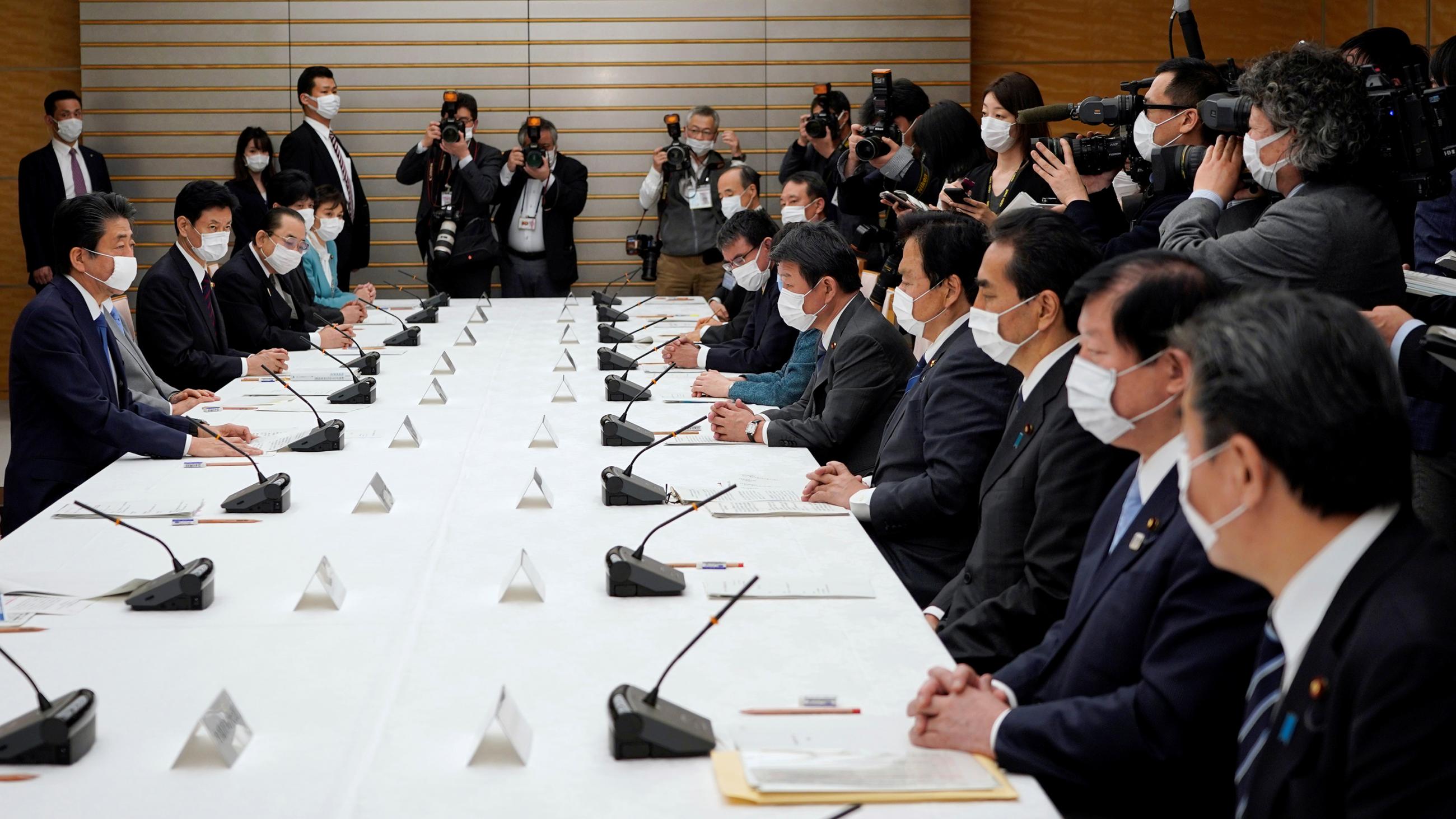 The photo shows the PM and a number of government ministers sitting around a large table. Reporters are seen at the edge of the frame snapping photos. Everyone in the picture is wearing a mask. 