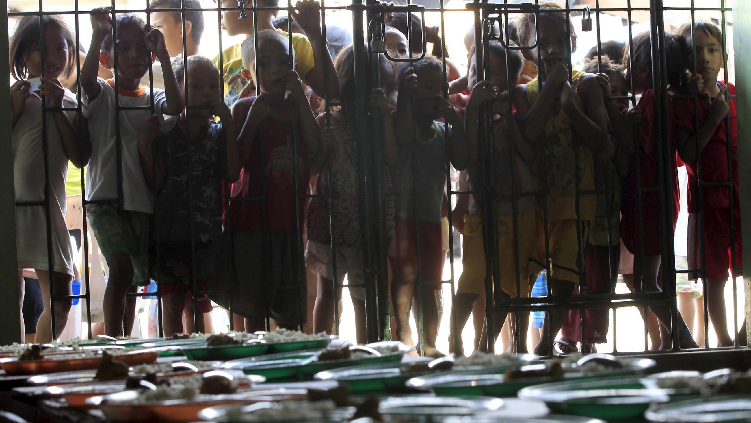 The photo shows children lined up along the bars keeping them from the interior of the space wherein the photographer is standing. In the foreground can be seen a table piled with individual meals. 