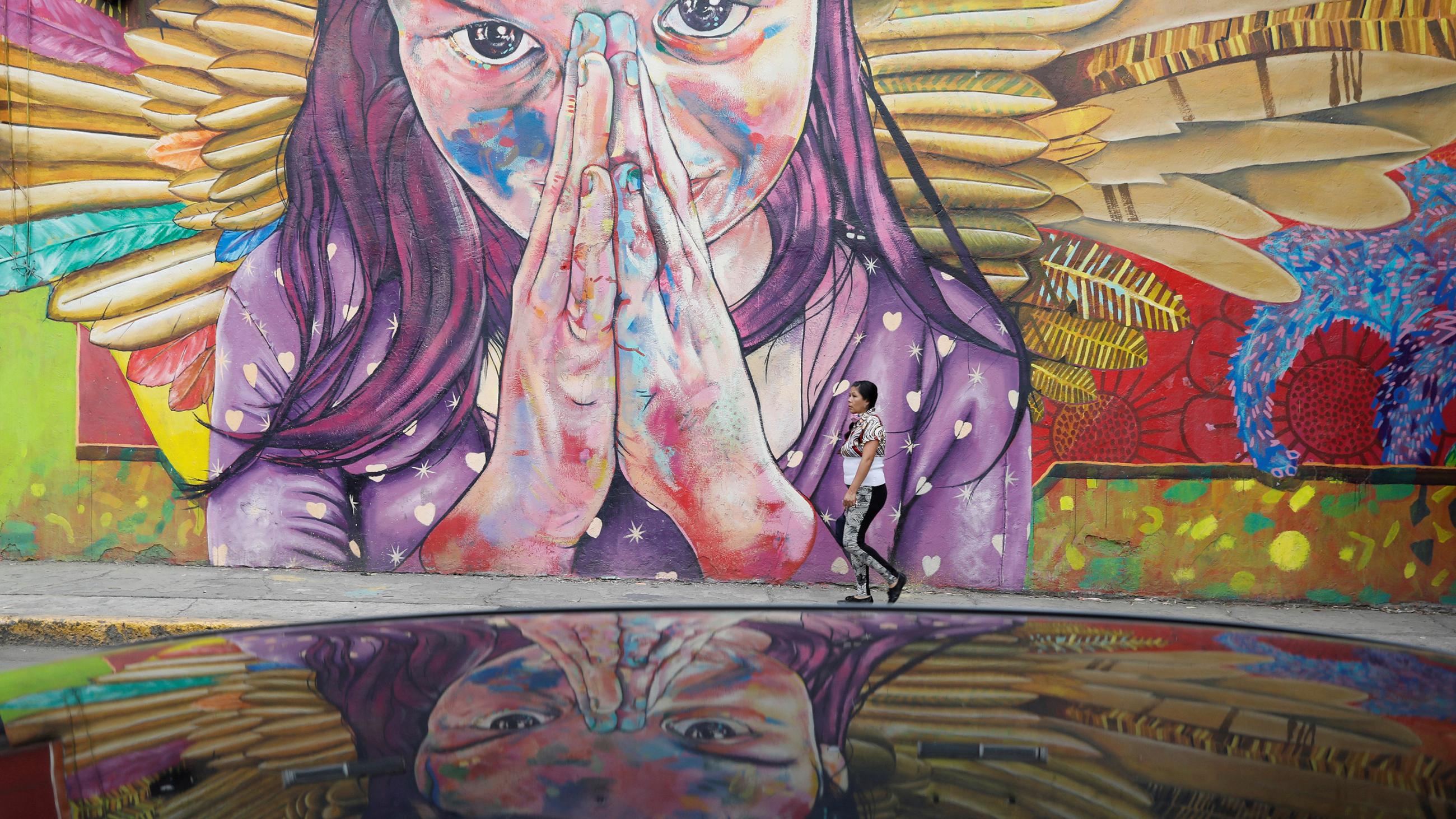 The photo shows an amazing mural on a wall of a woman with angel's wings and her hands together, as if in prayer. A woman walking in front of the mural demonstrates that the scale of the painting is massive. 