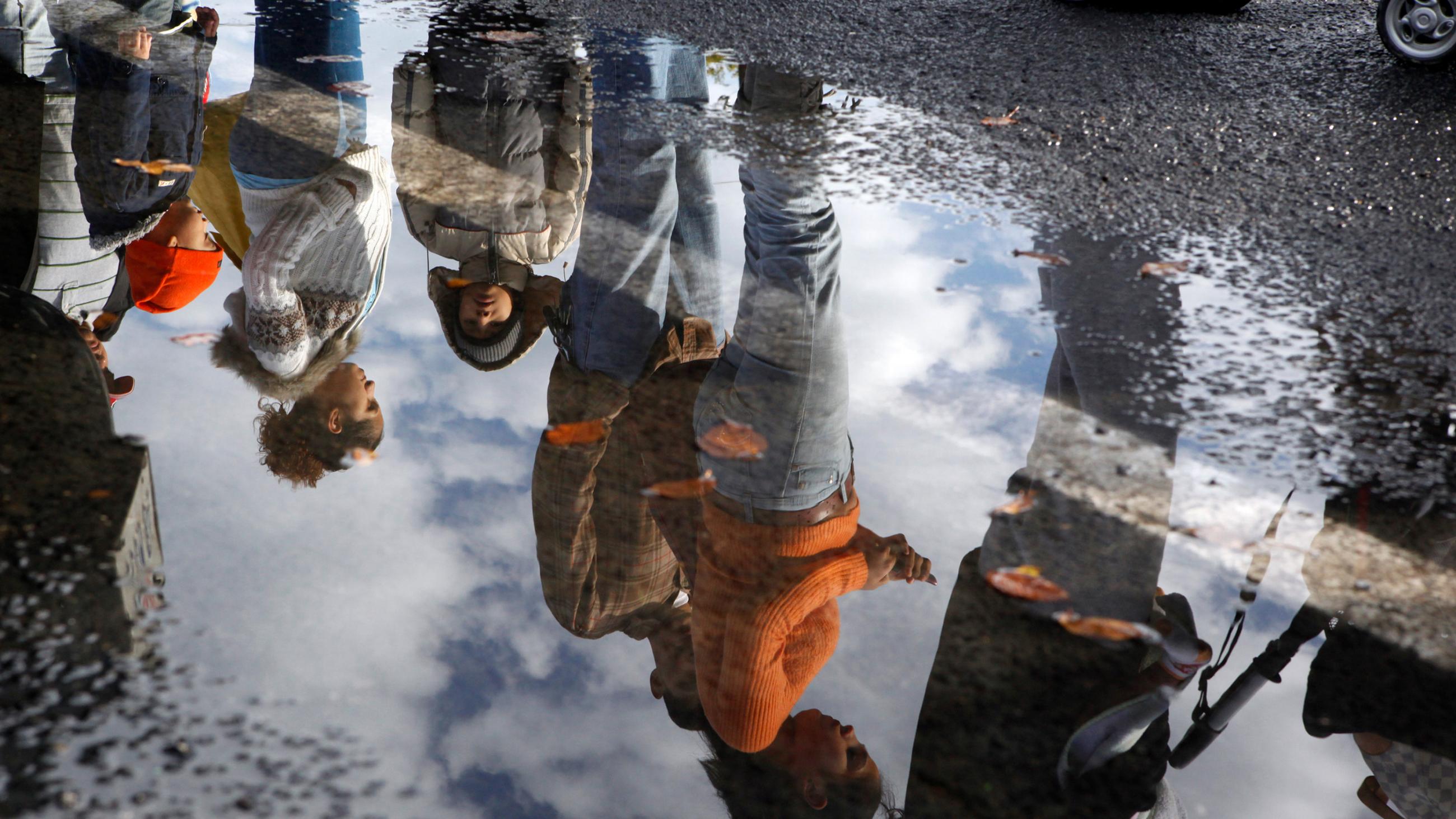 The photo shows a mud puddle with people standing in line reflected in the water. 