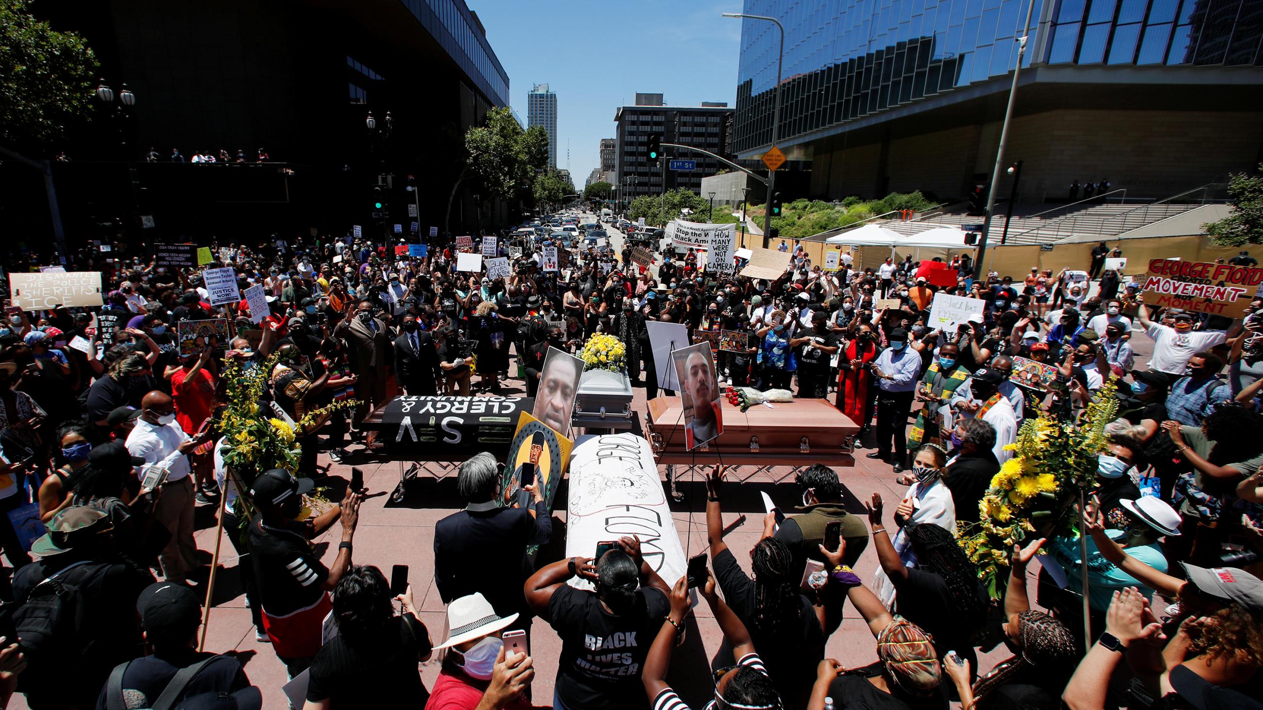 The photo shows a large crowd gathered around some coffins arranged in a cross-like formation. 