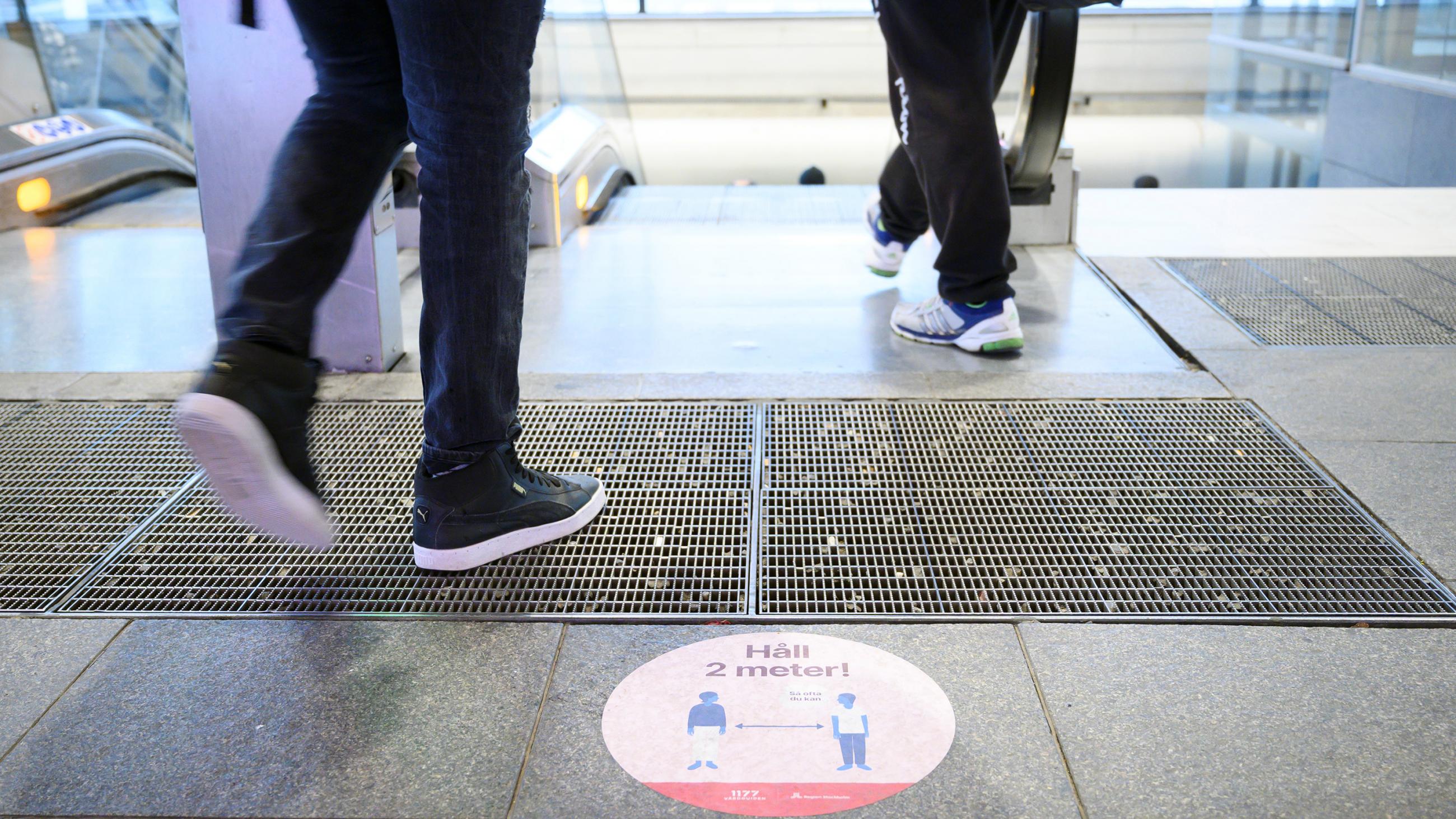 The photo shows feet walking onto an escalator above a sign warning of social distancing. 