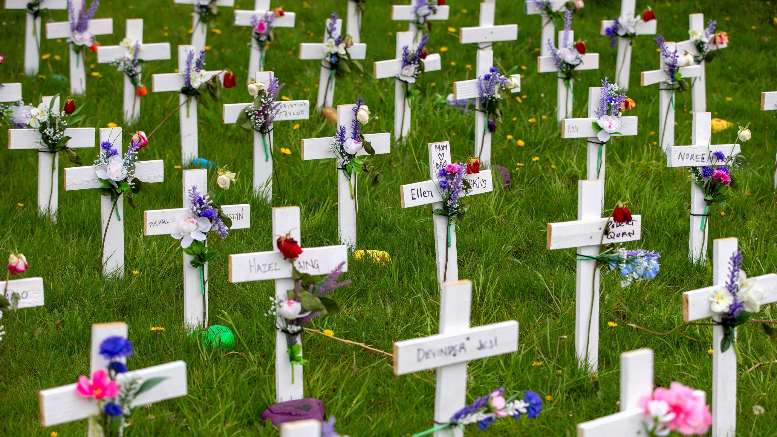 The photo shows a field of simple white wooden crosses, each with a hand-written name and a flower affixed to the front, in a grassy field. 