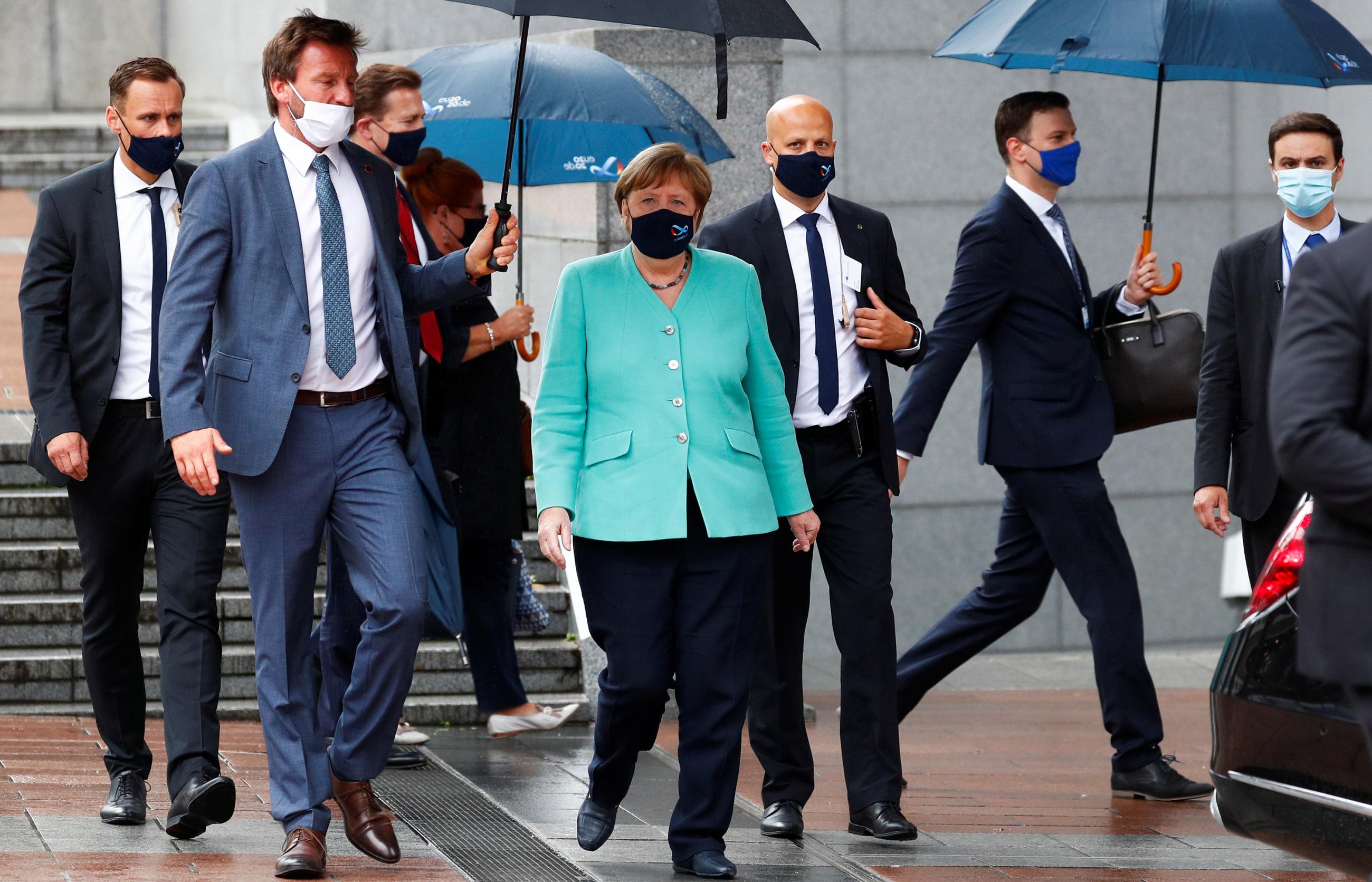 The photo shows Merkel walking toward the camera wearing a face mask amid a gaggle of men. She wears a pastel green blazer that stands out against their dark suits. One of her staff holds an umbrella out, shielding her. 