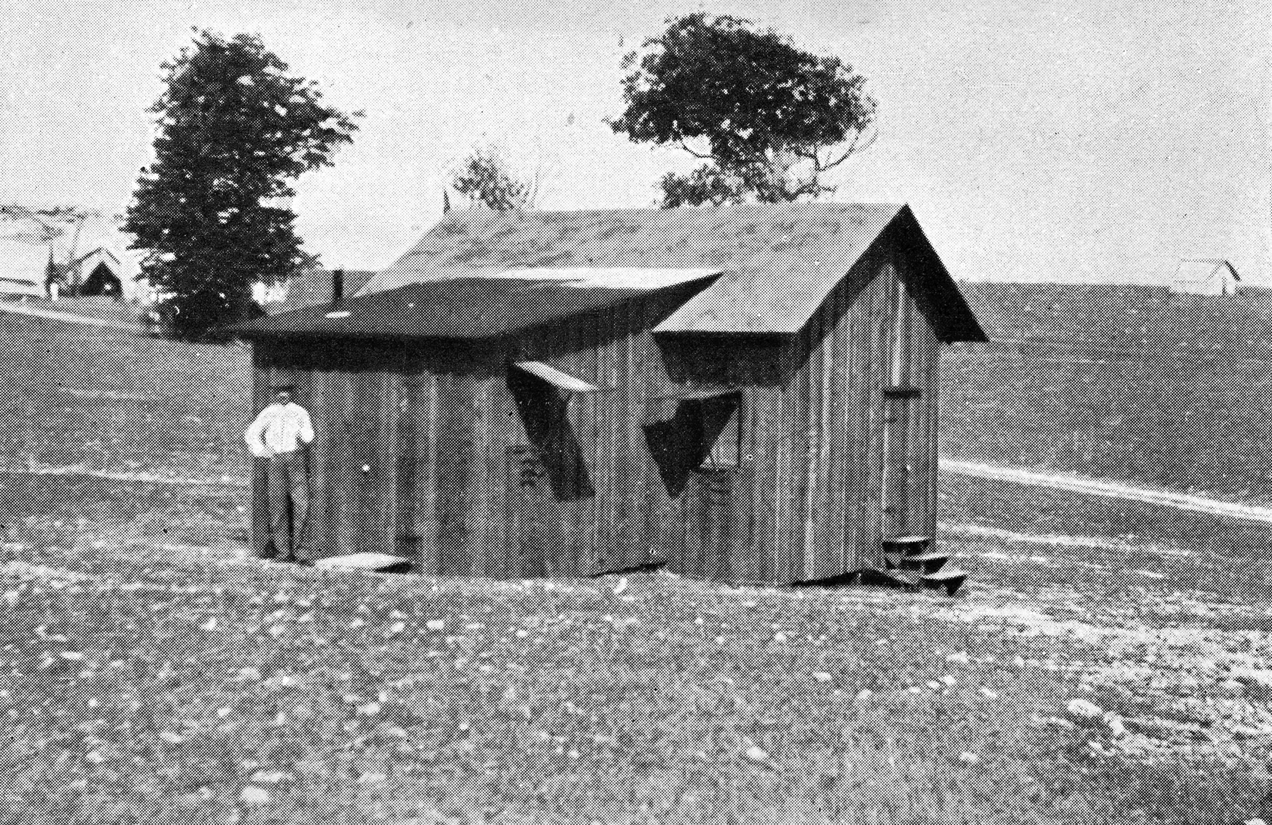 The historic photo shows a wooden hut of a building in a desolate setting with a man standing out front. 