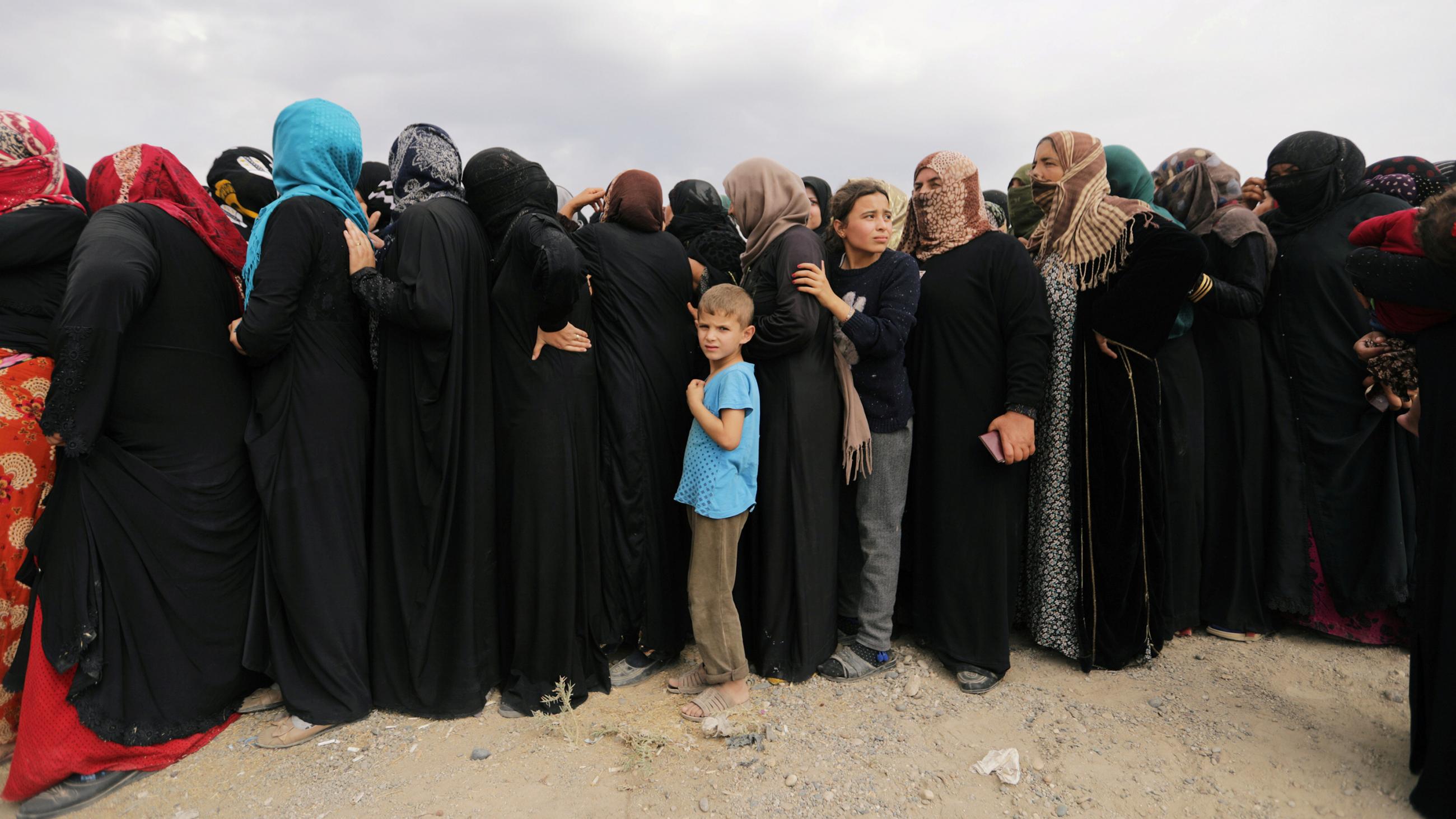 The photo shows a line of mostly women, all in black coverings, with a single boy in a light blue shirt in line with them. It is striking because his shirt stands out against a sea of black. 
