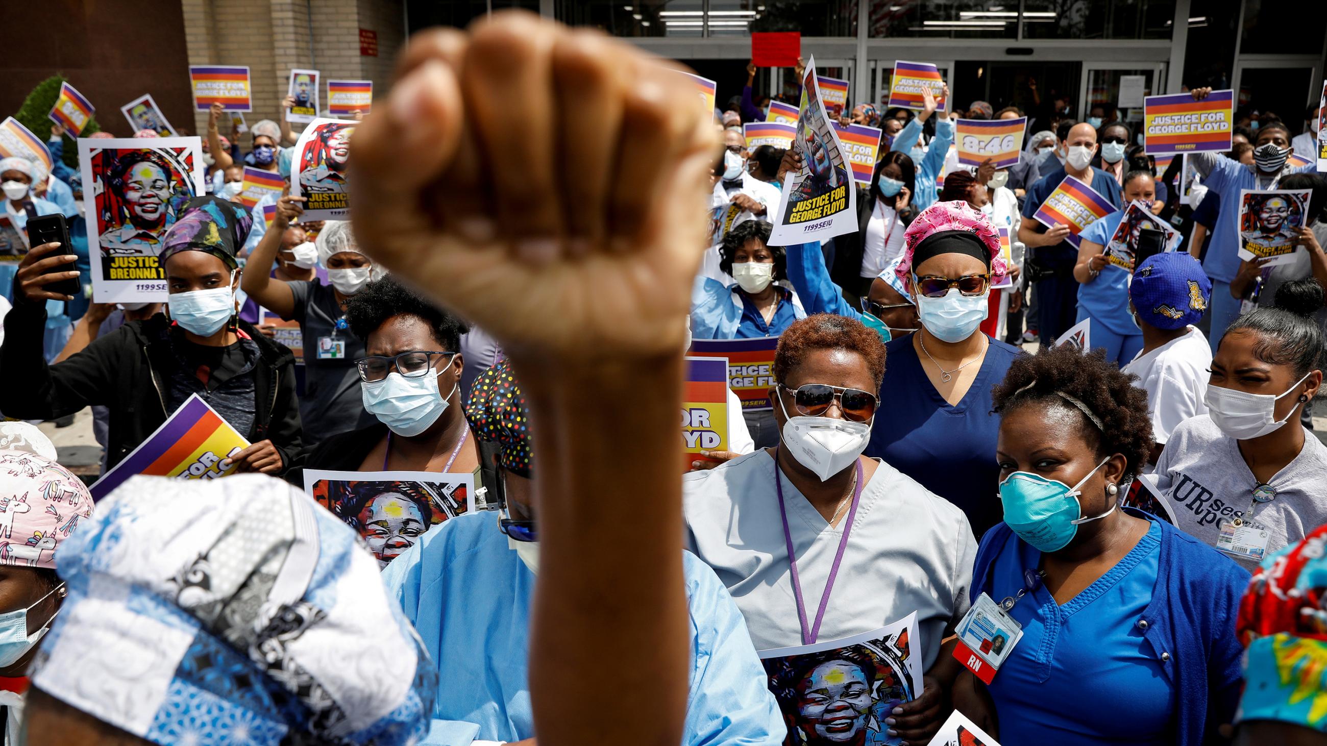 The photo shows people kneeling in protest while one protester holds a fist high in the camera's foreground. 