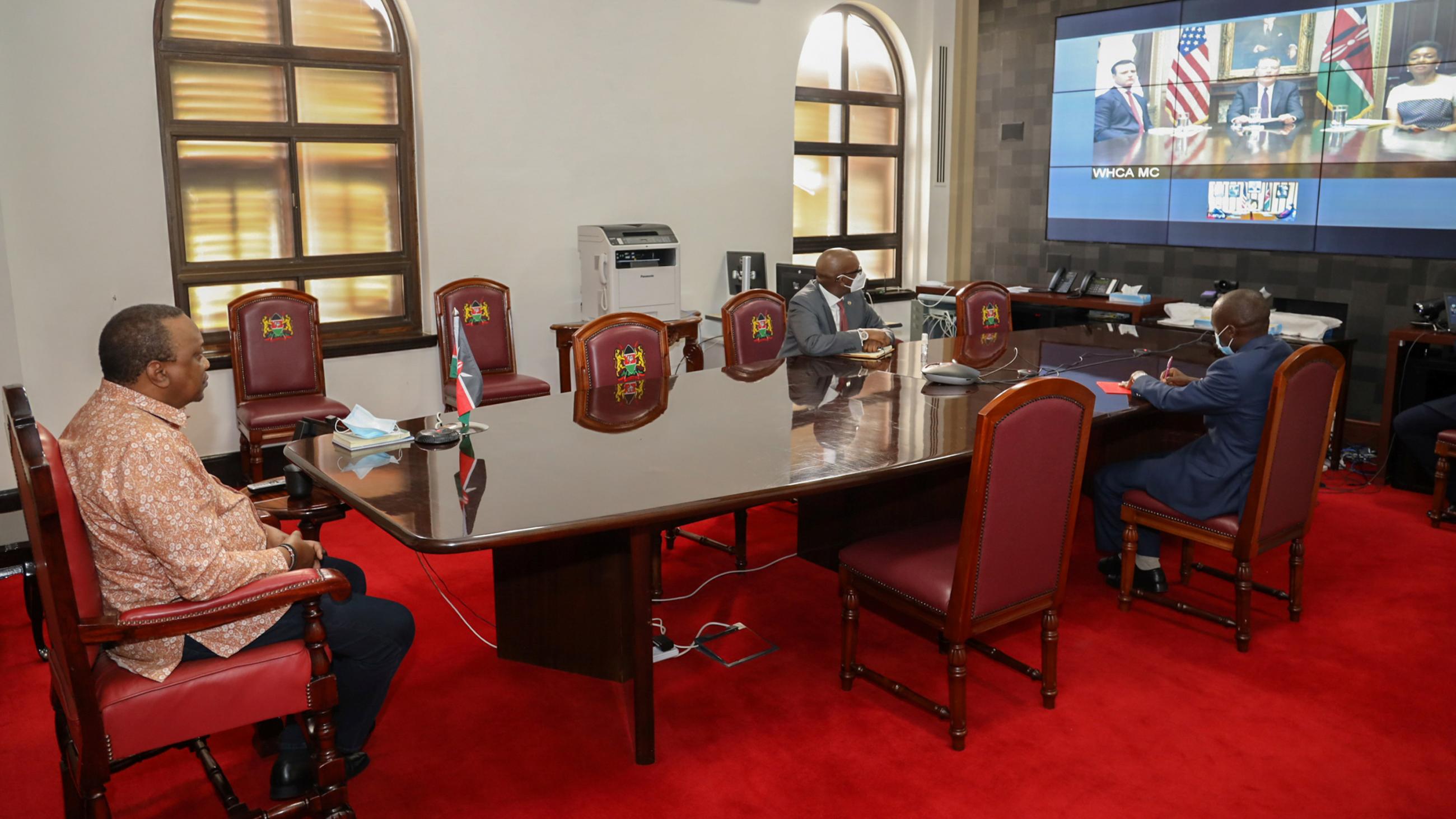 Photo shows the president in a room at a large table watching a large display screen across the room. 