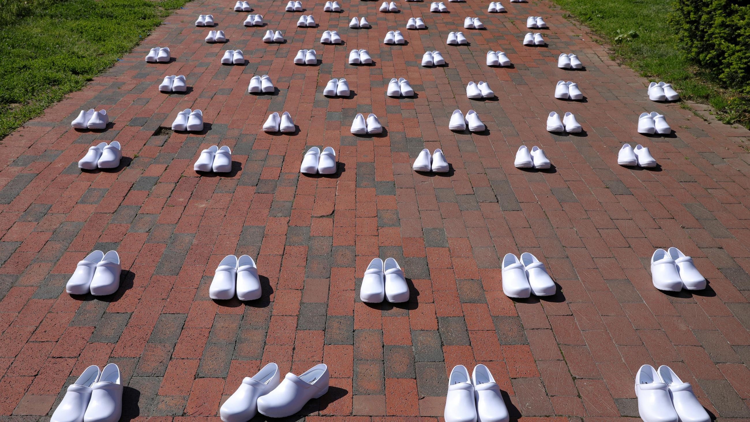The photo shows an array of white shoes lined up along a paved path. One pair in the front is slightly askew. 