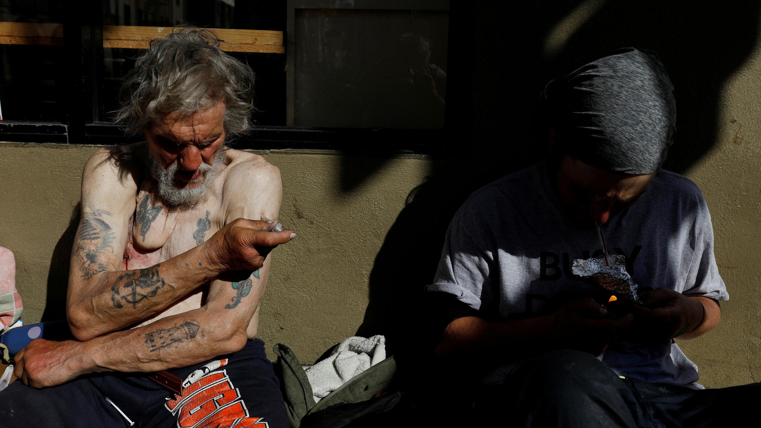 The photo shows a shirtless man with numerous tattoos injecting himself in the left arm. 